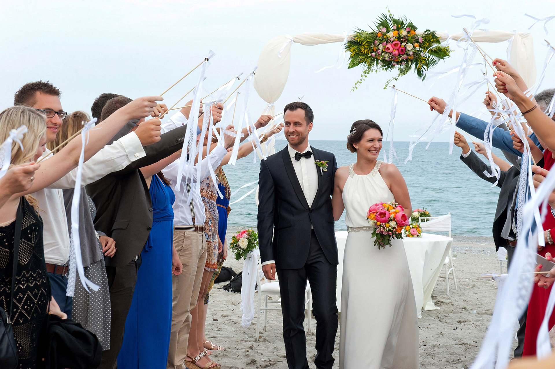 Το ζευγάρι χαμογελά καθώς περπατά ανάμεσα στους καλεσμένους με φόντο τη θάλασσα. The bride and groom smile while walking through the guests with the sea in the background.