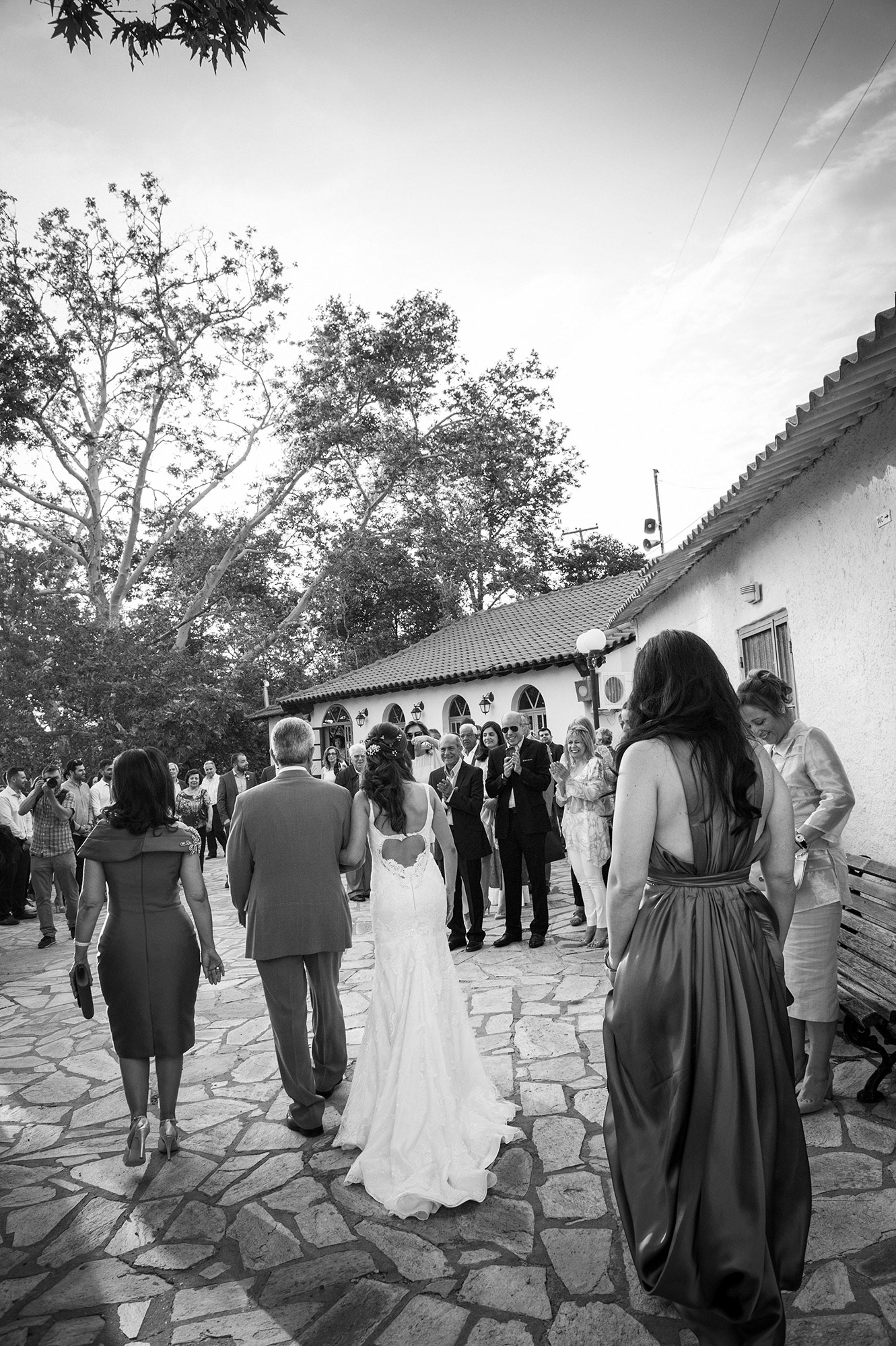 Η νύφη φτάνει στην εκκλησία πιασμένη από το χέρι του πατέρα της, με καλεσμένους να την υποδέχονται συγκινημένοι.    The bride arrives at the church holding her father’s arm, as guests welcome her with emotion.