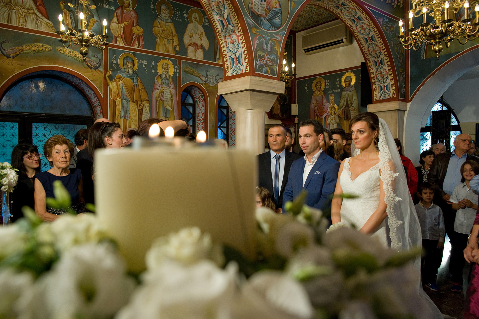 Γενικό πλάνο μέσα στην εκκλησία με θολή τη λαμπάδα στο πρώτο πλάνο και το ζευγάρι στο βάθος – καλλιτεχνική φωτογράφηση γάμου., General shot inside the church with the candle in the foreground blurred and the couple in the background - artistic wedding photography.