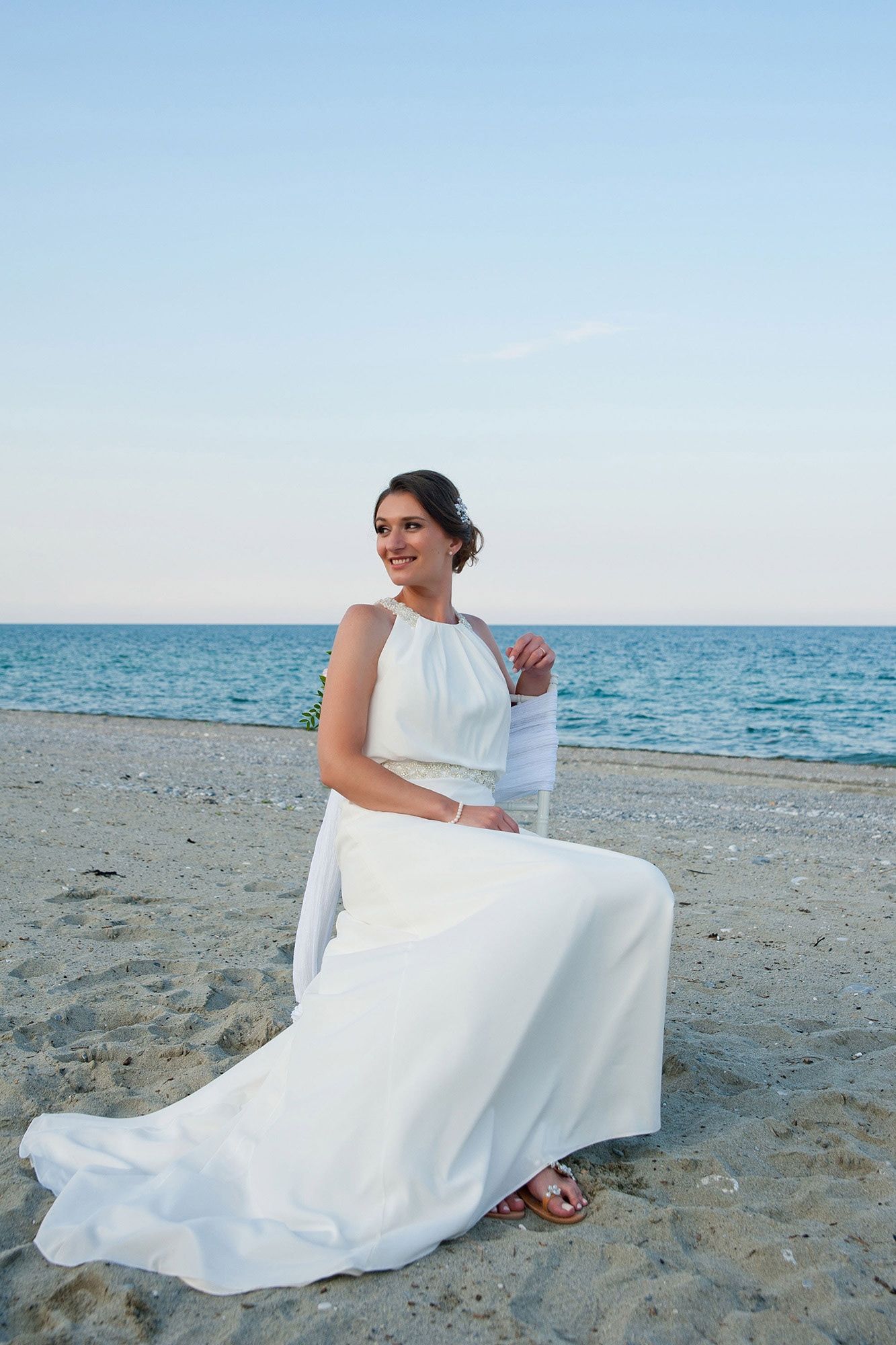 Η νύφη κάθεται στην καρέκλα στην παραλία, χαμογελώντας με φόντο τη θάλασσα.   The bride sits on a chair at the beach, smiling with the sea in the background.