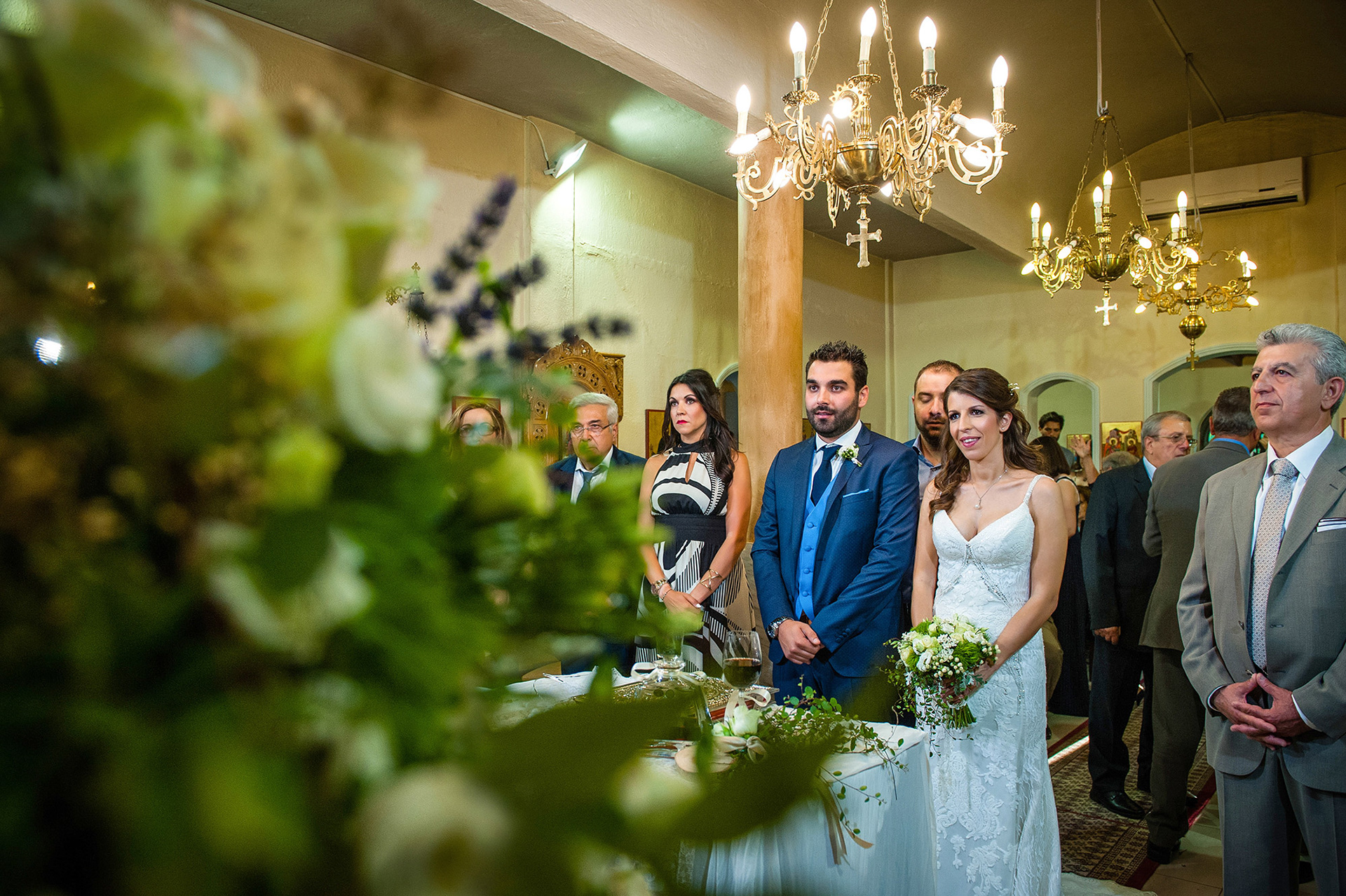 Ο γαμπρός και η νύφη στέκονται μπροστά στο ιερό της εκκλησίας κατά τη διάρκεια της τελετής, με βλέμματα γεμάτα χαρά.    The bride and groom stand before the altar during the ceremony, smiling with joy.