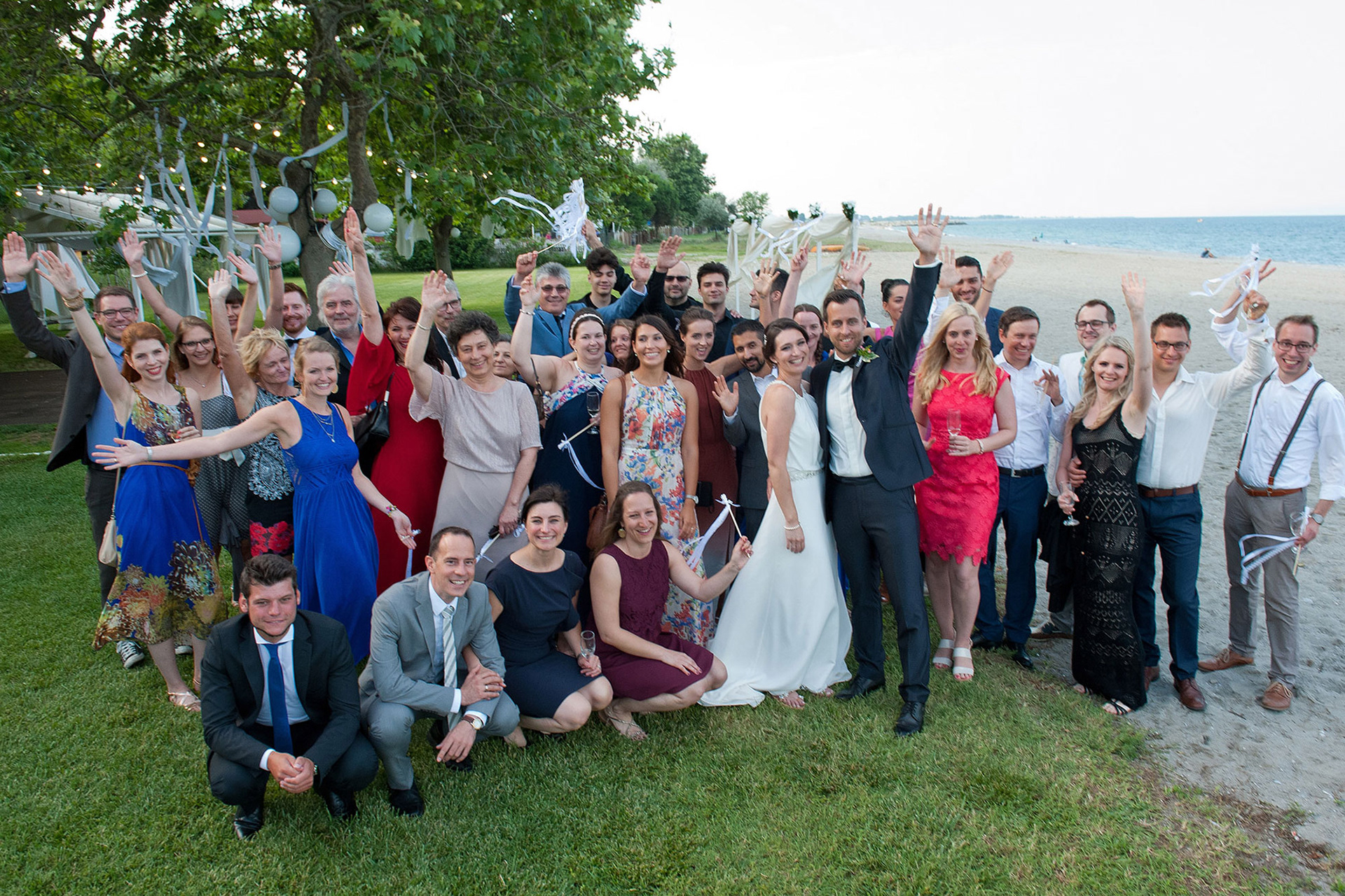 Ομαδική φωτογραφία του ζευγαριού με όλους τους καλεσμένους δίπλα στη θάλασσα.   Group photo of the couple with all the guests by the seaside.