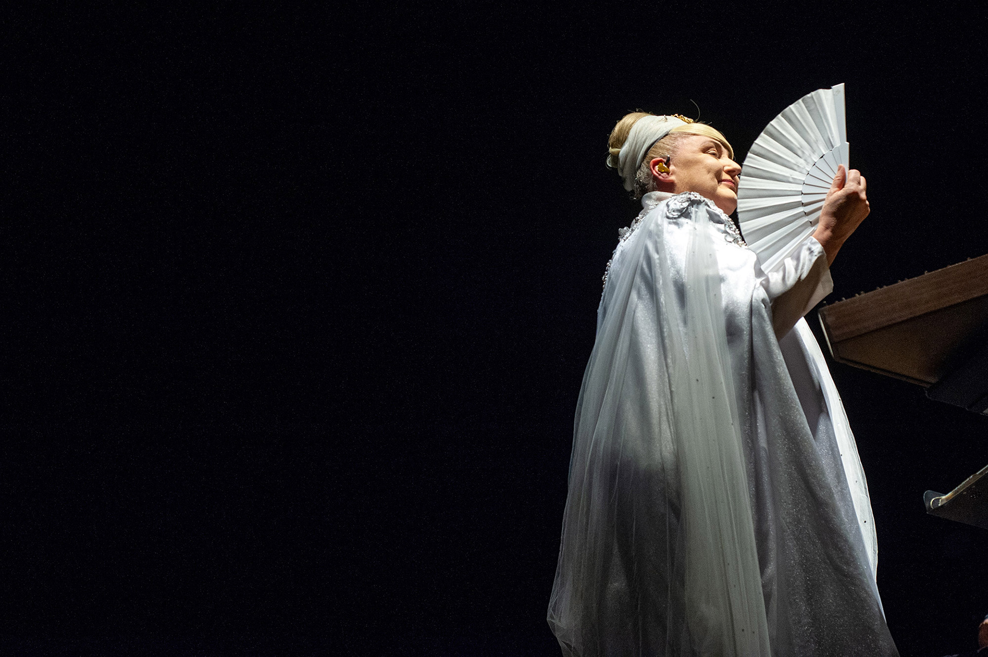 Καλλιτεχνική λήψη με τη Lisa Gerrard να κρατά μια λευκή βεντάλια, δημιουργώντας μια έντονη γεωμετρική αντίθεση με το μαύρο φόντο - Artistic shot of Lisa Gerrard holding a white fan, creating a sharp geometric contrast against the black backdrop