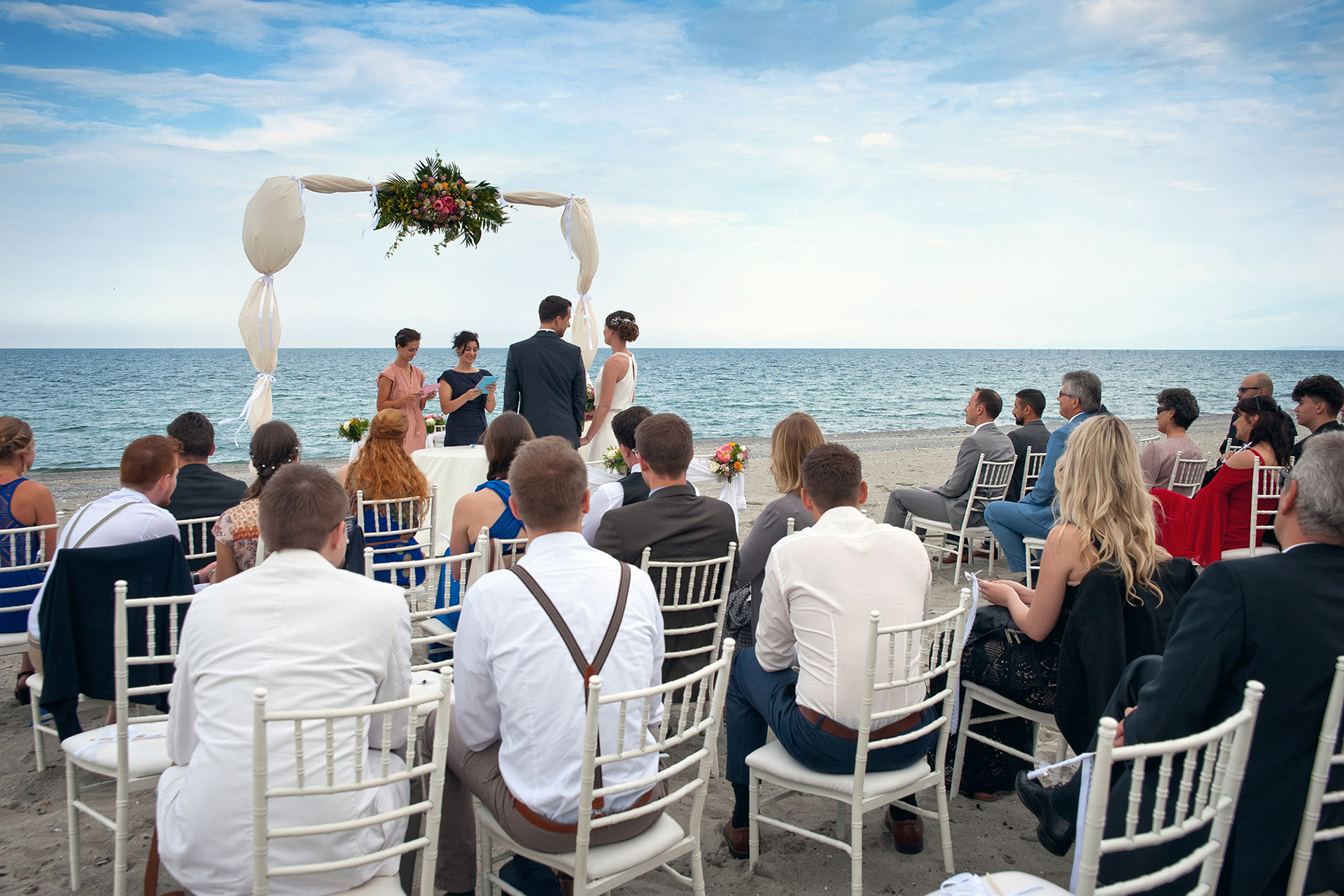 Η νύφη και ο γαμπρός κάτω από τον ανθοστόλιστο θόλο της τελετής με φόντο τη θάλασσα στο Λιτόχωρο.  The bride and groom standing beneath the floral arch with the sea in the background in Litochoro.