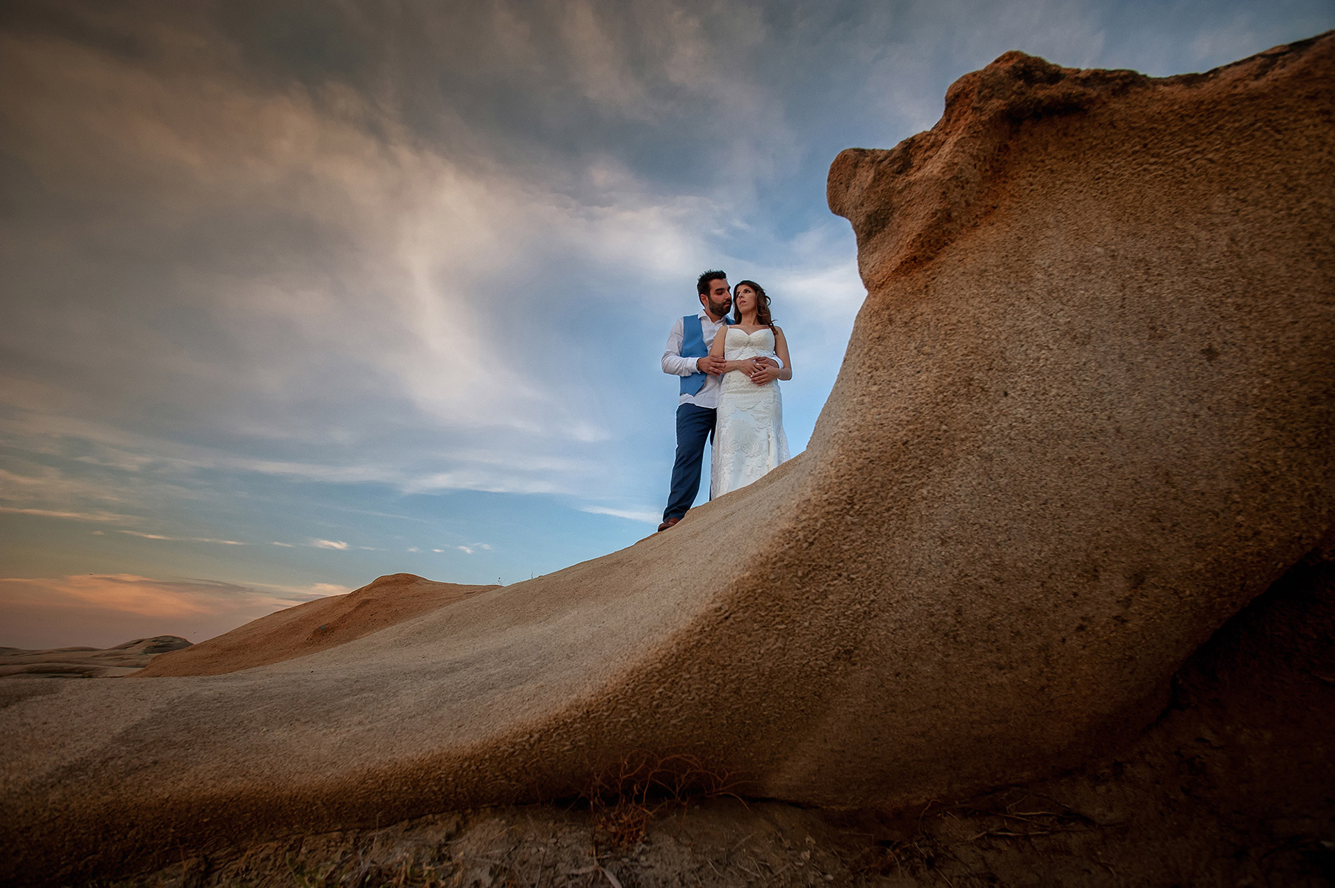 Το ζευγάρι αγκαλιά πάνω στους βράχους στη Σάρτη, κάτω από τον απογευματινό ουρανό, σε μια ρομαντική next day φωτογράφιση.    The couple embracing on the rocks in Sarti under the evening sky during a romantic next day wedding photoshoot.