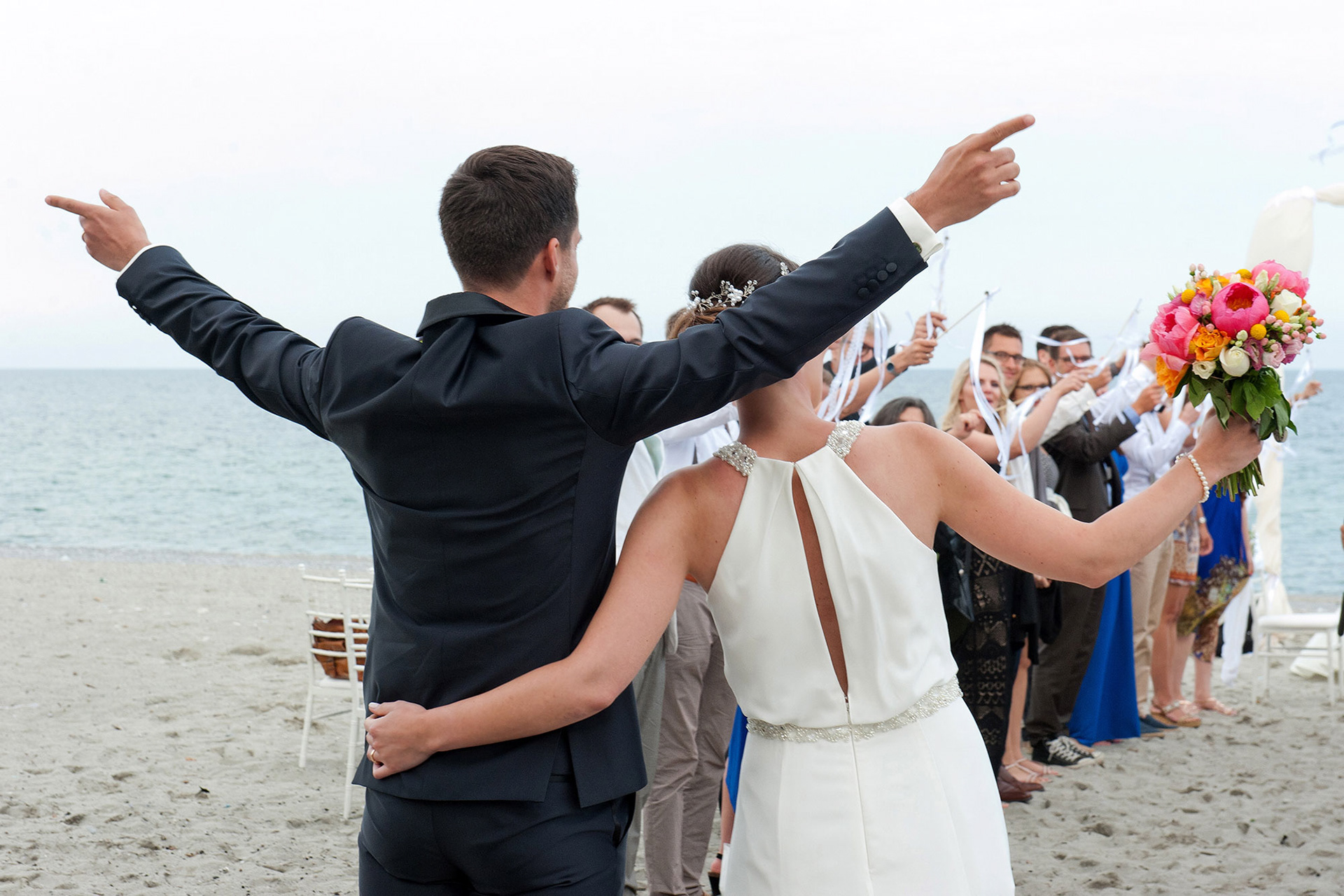 Το ζευγάρι με γυρισμένη την πλάτη κοιτά τους καλεσμένους σηκώνοντας τα χέρια με χαρά. The couple faces away from the camera, raising their arms joyfully toward the guests.