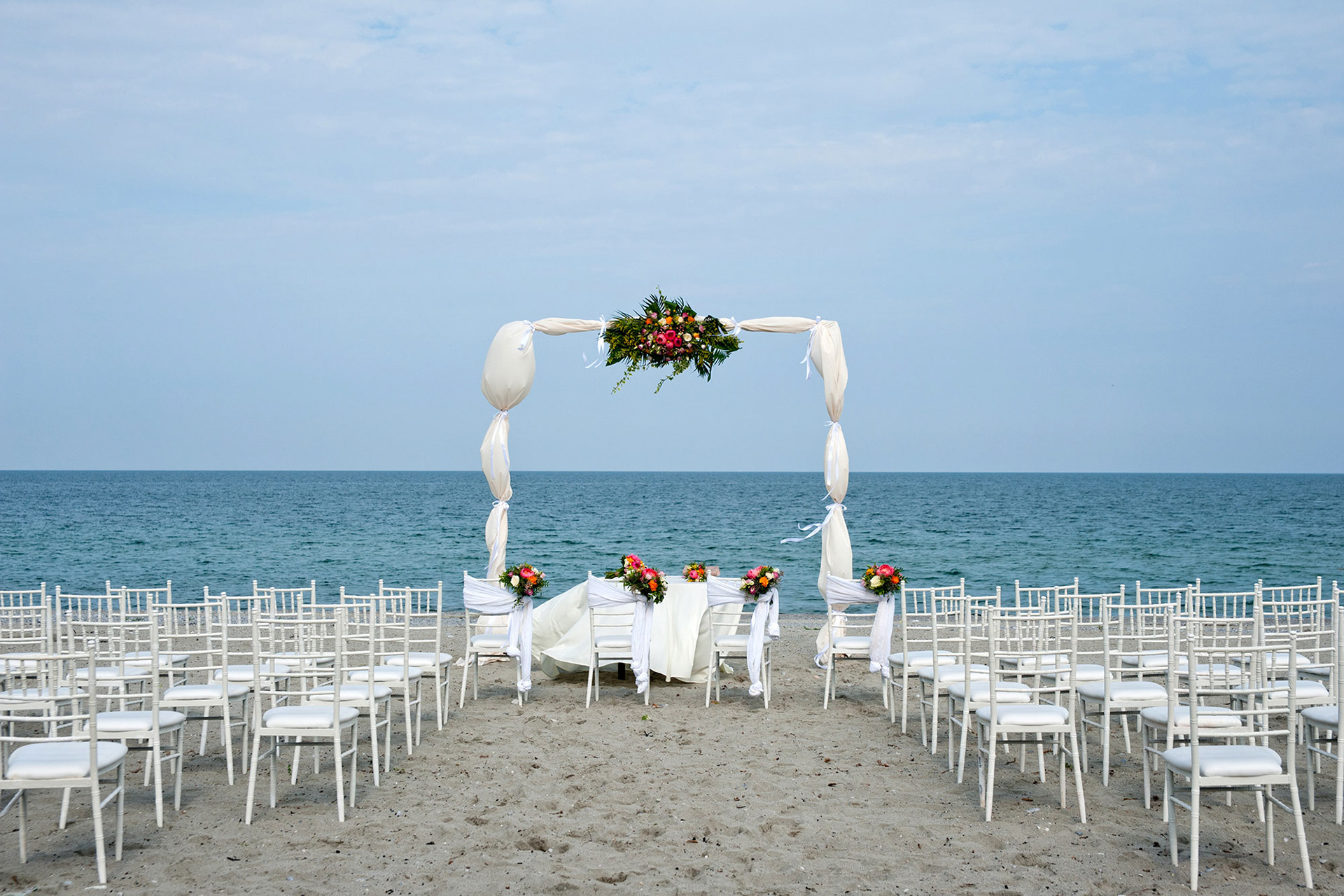 Η αψίδα του γάμου στολισμένη με λευκά υφάσματα και λουλούδια δίπλα στη θάλασσα.   The wedding arch draped in white fabric and flowers by the seaside.