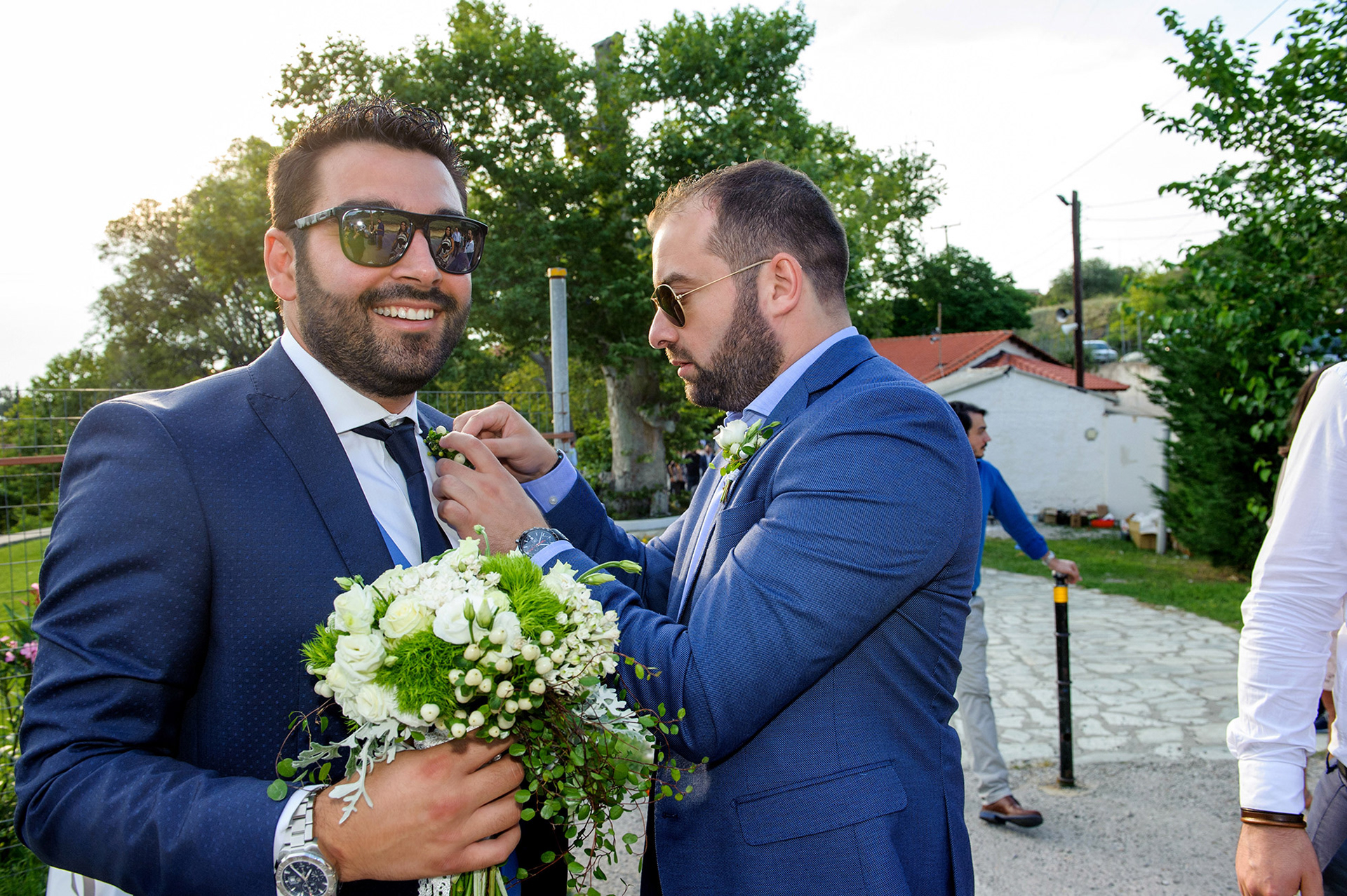 Ο κουμπάρος βοηθά τον γαμπρό να τοποθετήσει το boutonnière στο σακάκι του, χαμογελαστή στιγμή φιλίας. The best man helping the groom adjust his boutonnière, cheerful friendship moment before the ceremony.