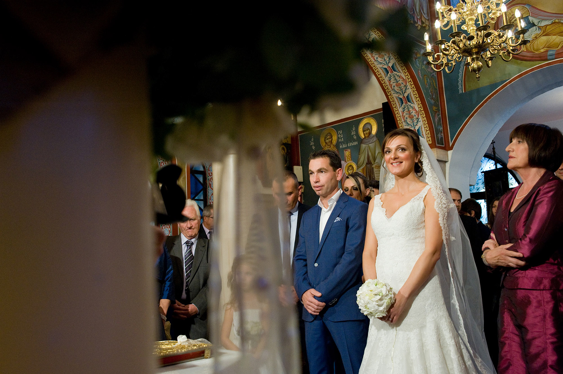 Γενικό πλάνο μέσα στην εκκλησία με θολό στολισμό στο πρώτο πλάνο και το ζευγάρι στο κέντρο – καλλιτεχνική φωτογράφηση γάμου., General shot inside the church with blurred decorations in the foreground and the couple in the center - artistic wedding photography.