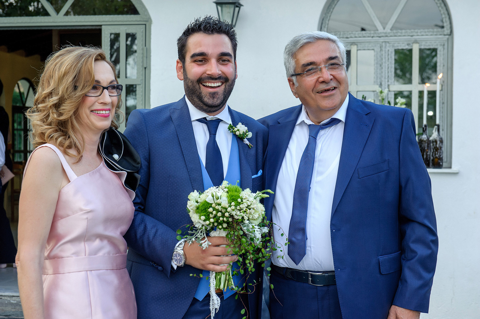 Ο γαμπρός φωτογραφίζεται χαμογελαστός με τους γονείς του έξω από την εκκλησία.      The groom poses happily with his parents outside the church before the ceremony.
