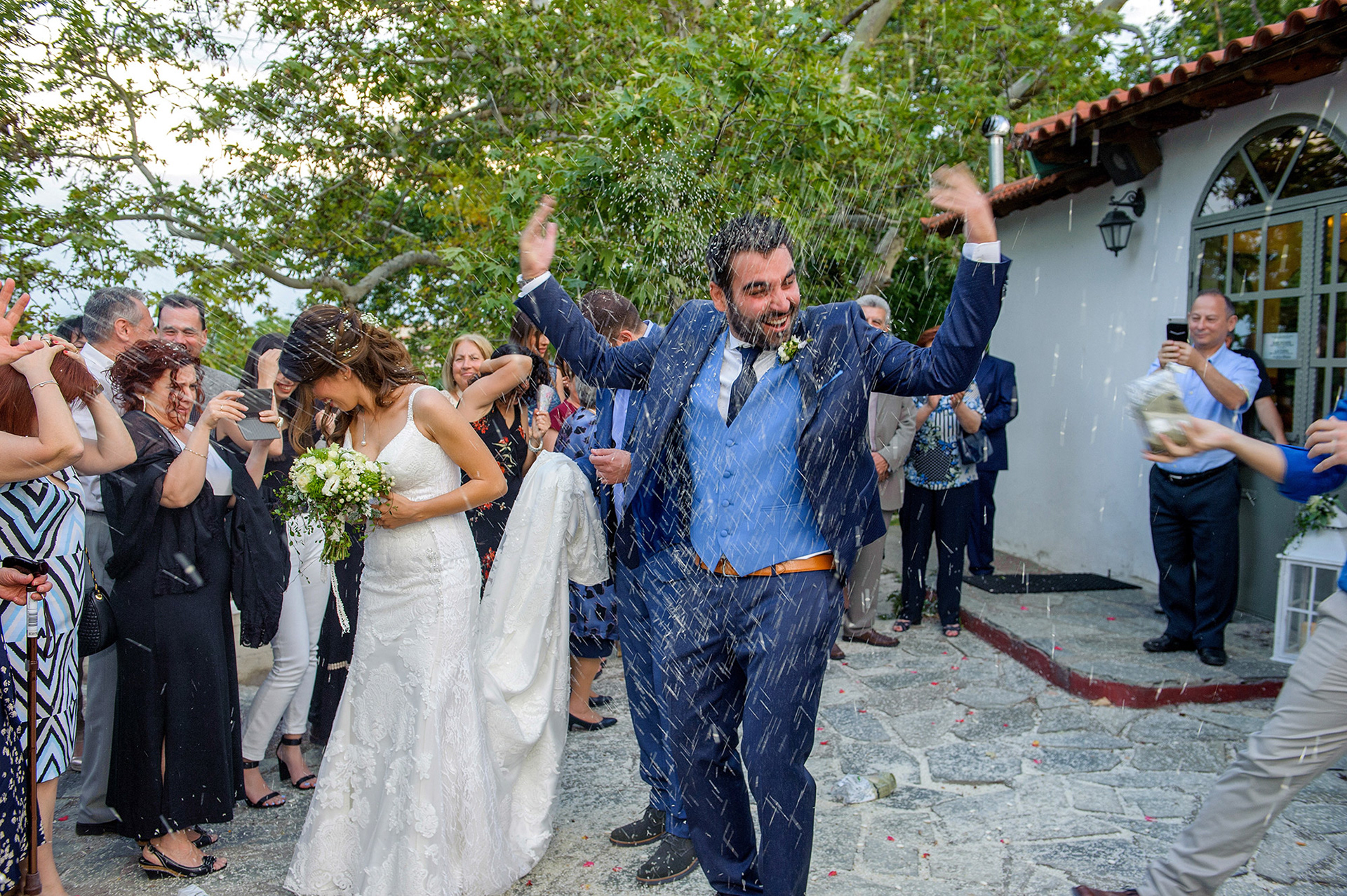 Το ζευγάρι γελά καθώς οι καλεσμένοι ρίχνουν ρύζι μετά το μυστήριο του γάμου.     The couple laughs joyfully as guests throw rice after the wedding ceremony.