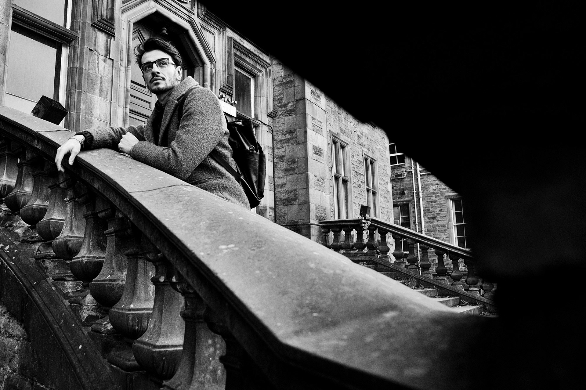 Μια ασπρόμαυρη fine art φωτογραφία ανδρικού πορτρέτου σε εξωτερικό χώρο από τη Βούλα Γκώτη, που αποτυπώνει έναν άνδρα με παλτό και γυαλιά σε μια ατμοσφαιρική στιγμή στις σκάλες ενός ιστορικού κτιρίου. | A moody black and white fine art outdoor portrait by photographer Voula Gkoti, capturing a man in a coat and glasses on the stone stairs of a historic building.
