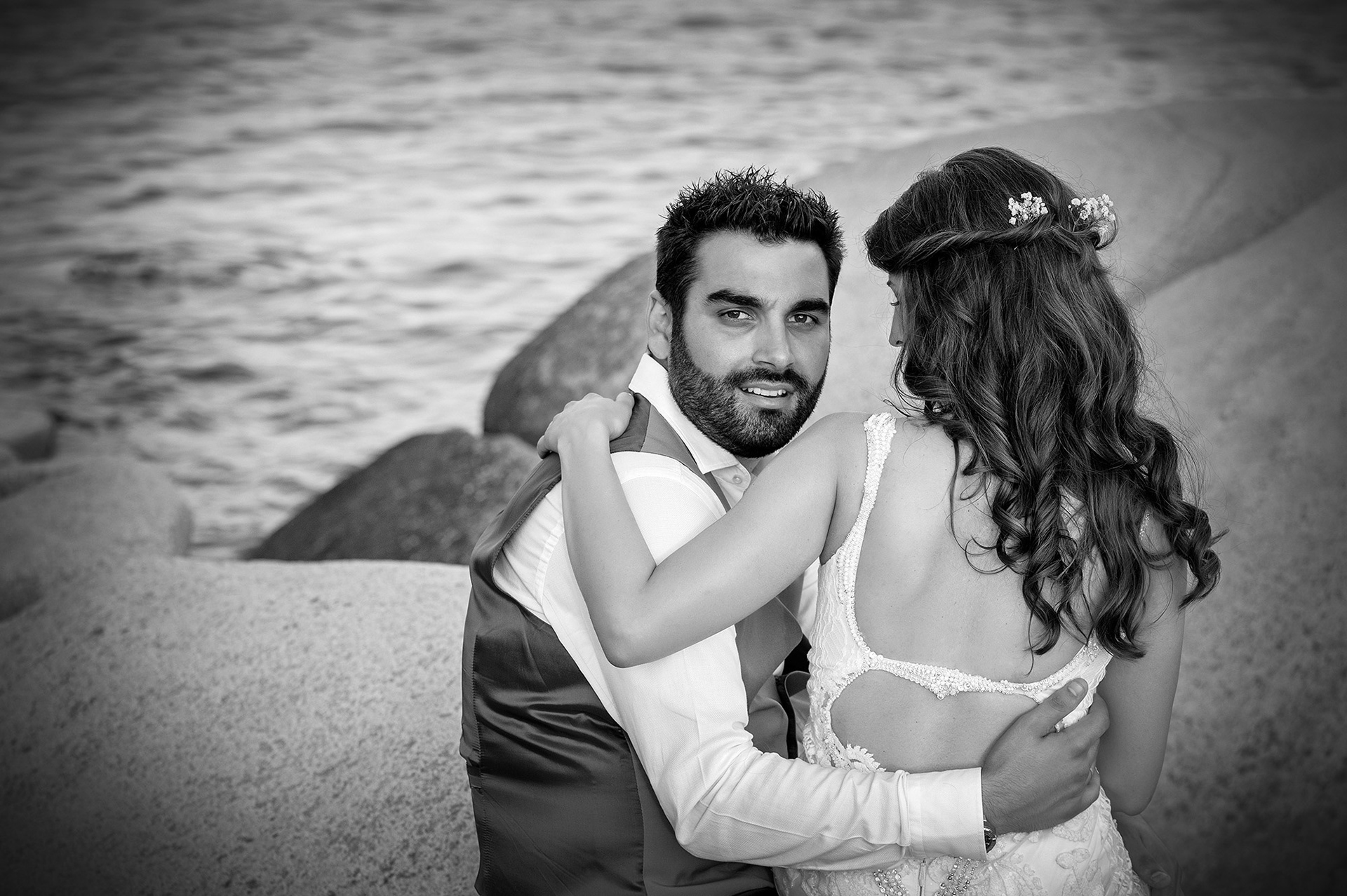 Ο γαμπρός κοιτάζει την κάμερα ενώ η νύφη τον αγκαλιάζει, με φόντο τη θάλασσα και τα βράχια.    The groom looks at the camera as the bride embraces him, with the sea and rocks in the background.