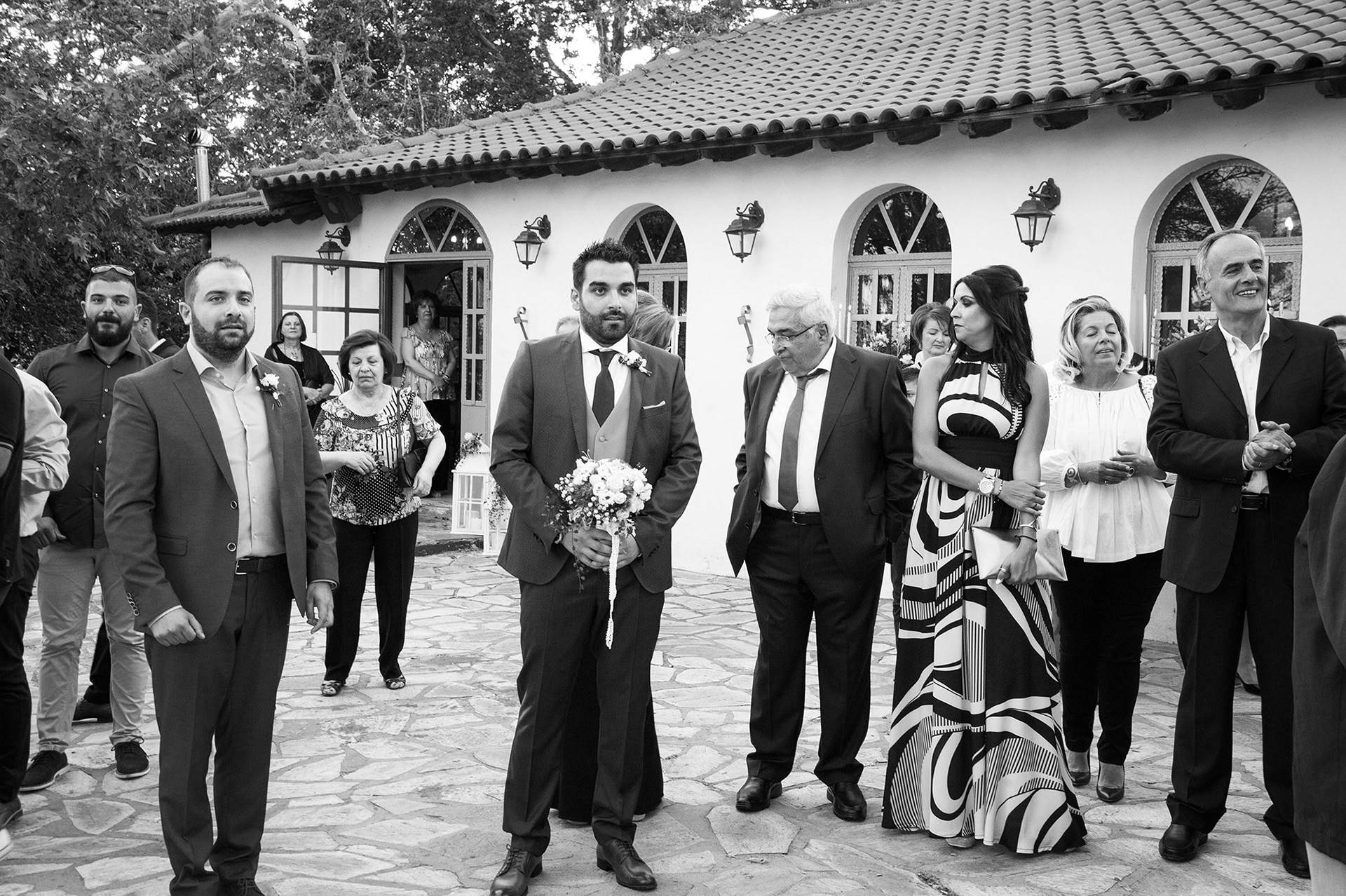 Ο γαμπρός περιμένει τη νύφη έξω από την εκκλησία, περιτριγυρισμένος από συγγενείς και φίλους.     The groom waits for the bride outside the church, surrounded by family and friends.