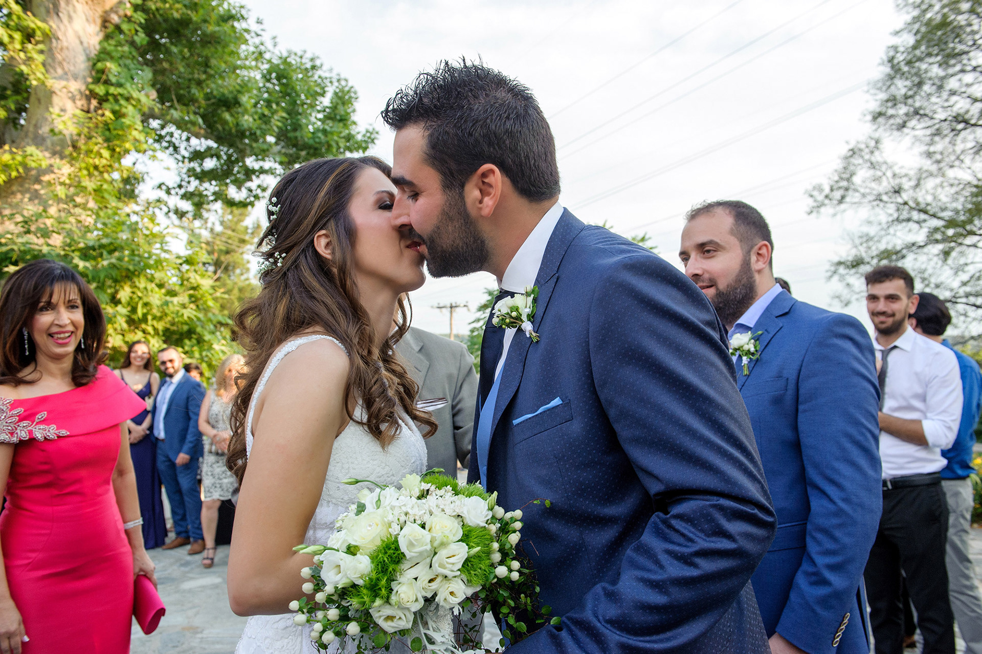 Ο γαμπρός και η νύφη μοιράζονται το πρώτο τους φιλί έξω από την εκκλησία, μέσα σε χειροκροτήματα και χαμόγελα.    The groom and bride share their first kiss outside the church, surrounded by smiles and applause.