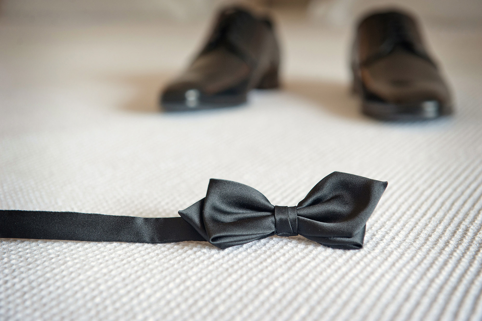 Μαύρο παπιγιόν και παπούτσια του γαμπρού τοποθετημένα στο δωμάτιο προετοιμασίας.   Black bow tie and groom’s shoes placed on the preparation room floor.