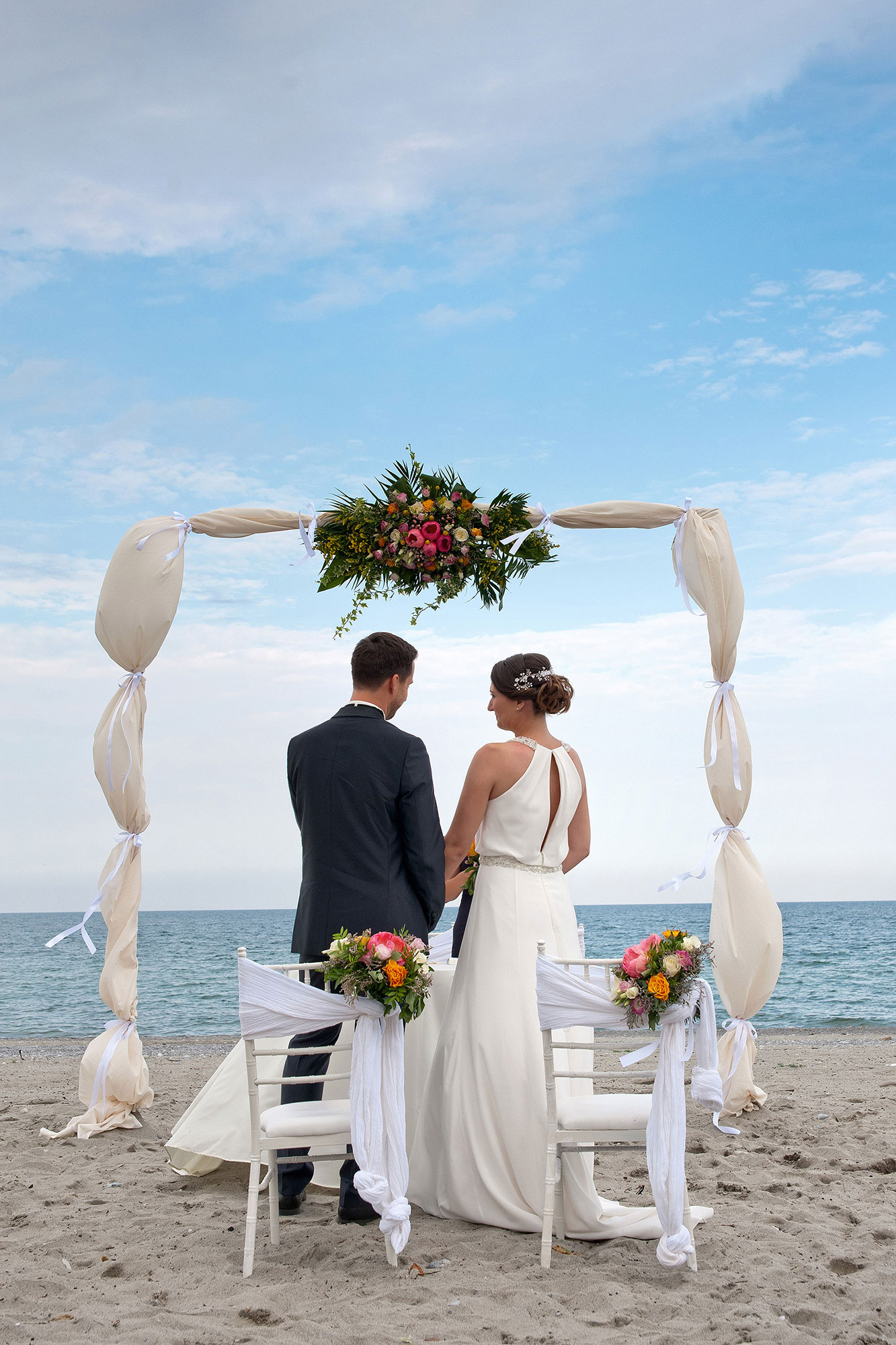 Η νύφη και ο γαμπρός κάτω από τον ανθοστόλιστο θόλο της τελετής με φόντο τη θάλασσα στο Λιτόχωρο.   The bride and groom standing beneath the floral arch with the sea in the background in Litochoro.