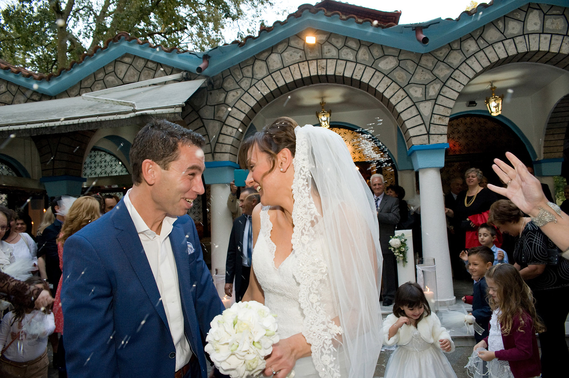 Το ζευγάρι έξω από την εκκλησία ενώ φίλοι και συγγενείς τους ραίνουν με ρύζι κοιτάζονται και γελάνε– φωτογράφηση γάμου., The couple outside the church while friends and relatives shower them with rice, looking at each other and laughing – wedding photography.