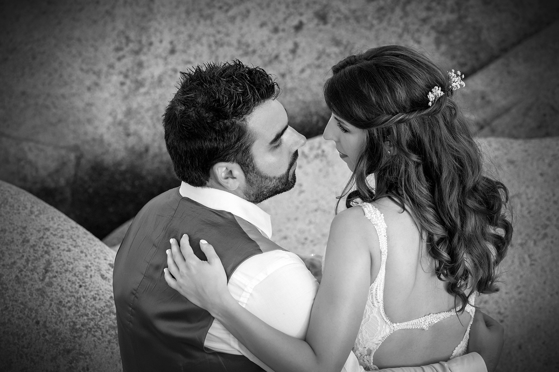 Ρομαντική στιγμή όπου η νύφη και ο γαμπρός κοιτάζονται στα μάτια ανάμεσα στα βράχια.    Romantic moment as the bride and groom look into each other’s eyes among the seaside rocks.