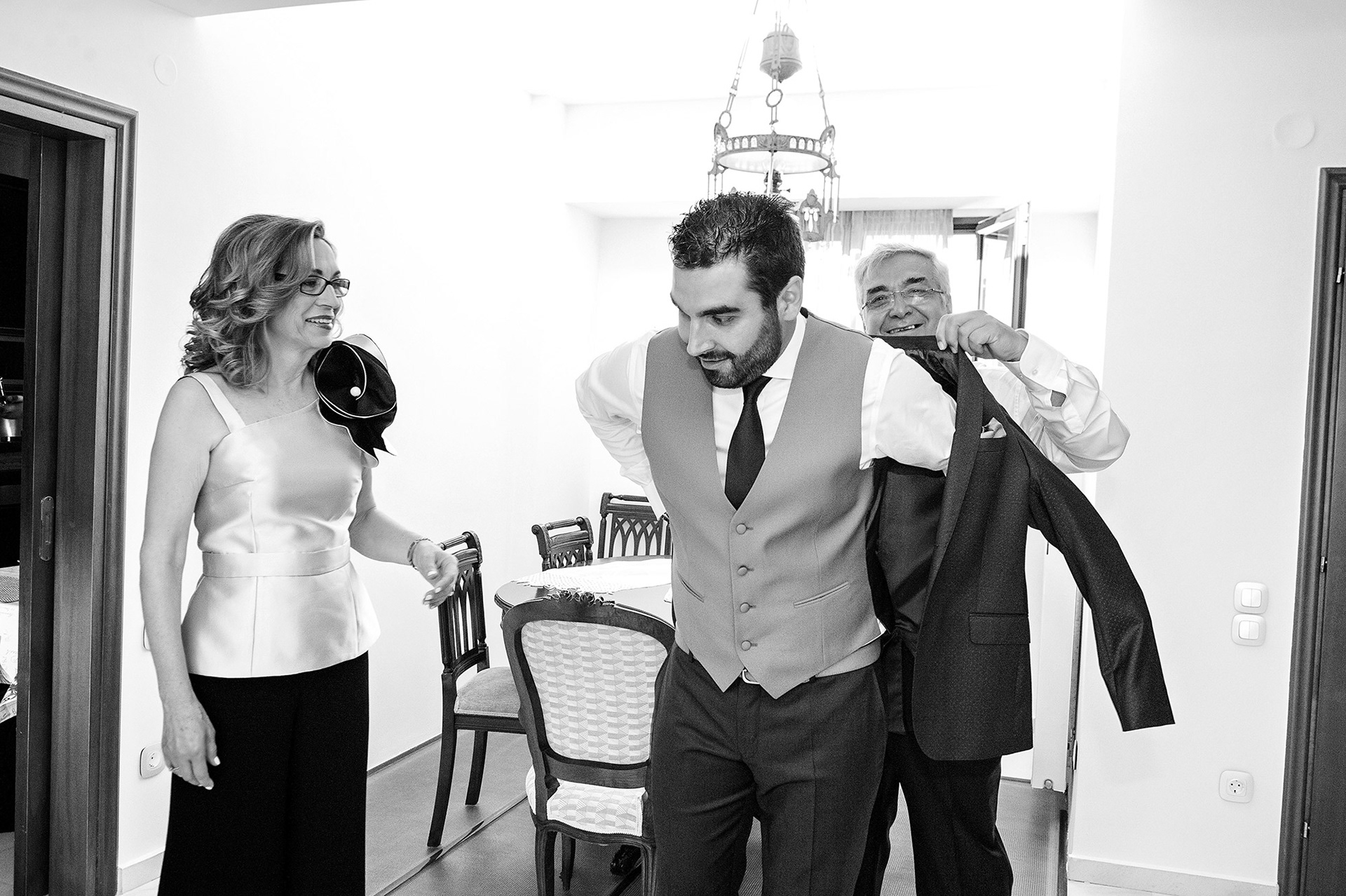 Οι γονείς βοηθούν τον γαμπρό να φορέσει το σακάκι του, τρυφερή στιγμή οικογενειακής προετοιμασίας.     Groom’s parents helping him with his jacket, tender family moment before the wedding.