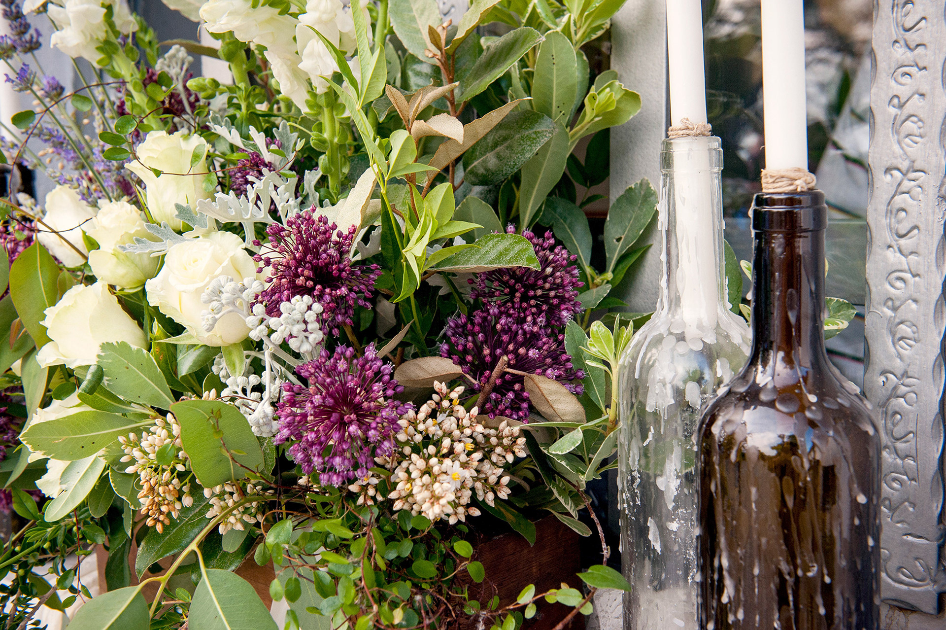 Ανθοσύνθεση με λευκά τριαντάφυλλα, μοβ άνθη και κεριά σε γυάλινες μπουκάλες.    Floral arrangement with white roses, purple blooms, and candles in glass bottles.