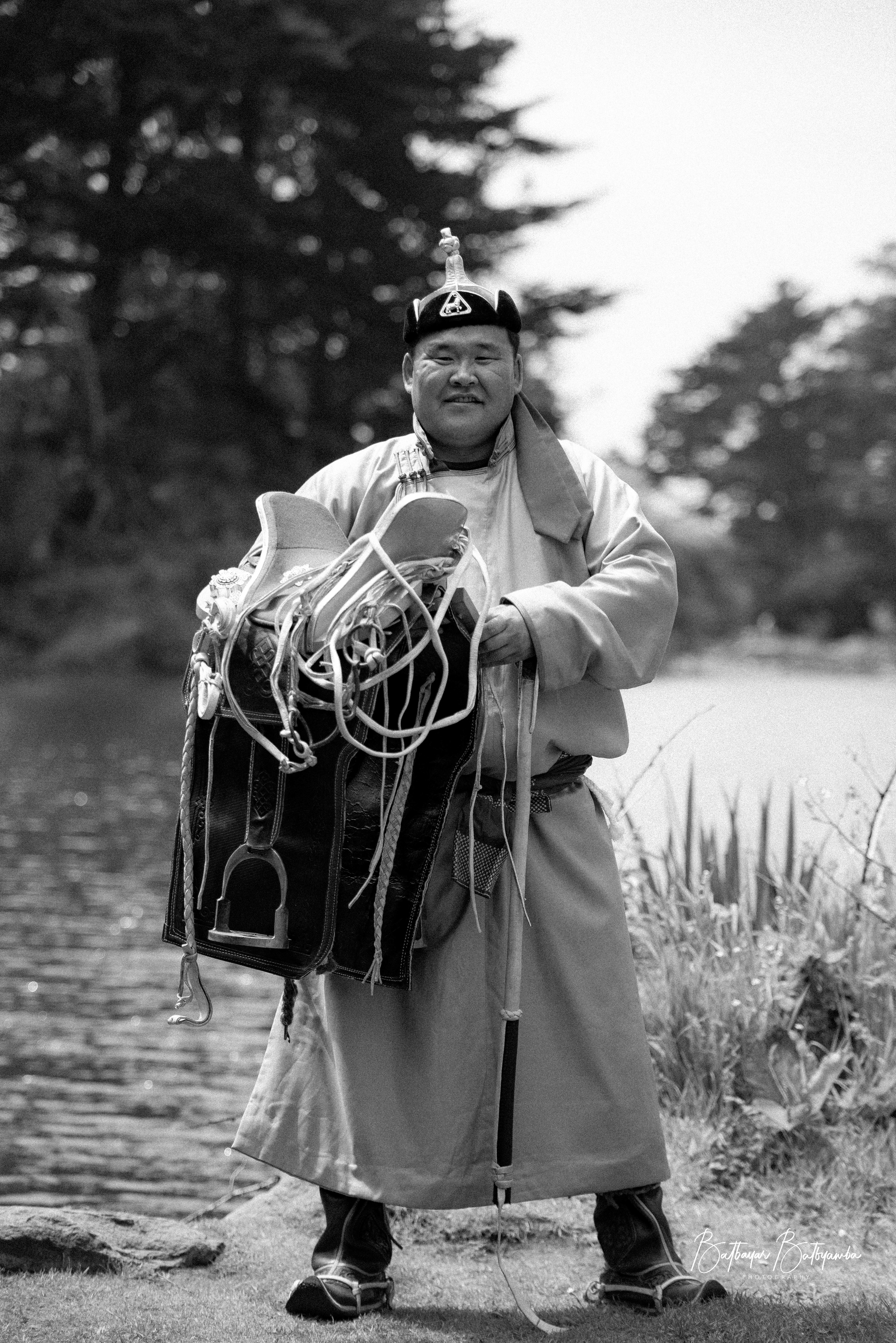 **Sumiin Zaan Chiinbaatar** is holding a **Mongolian emeel**, a traditional saddle renowned for its unique craftsmanship and cultural significance. The Mongolian emeel is meticulously handcrafted from wood, leather, and metal, often adorned with intricate carvings and silver or gold embellishments. Designed for durability and comfort, it reflects the nomadic lifestyle of Mongolia, where horses are central to daily life. The emeel not only serves as a functional tool for horseback riding but also symbolizes status, heritage, and the skilled artistry passed down through generations.