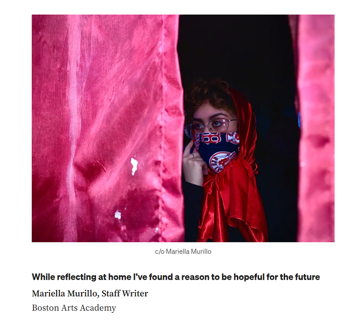 A screenshot of one of Mariella Murillo's perspective pieces. In the image, she is wearing a glasses, a red head scarf, a Boston Red Sox-face masks, and glasses, partially hidden behind a red curtain. Underneath the image is the headline "While reflecting at home I've found a reason to be hopeful for the future", byline "Mariella Murillo, Staff Writer," and her affiliation "Boston Arts Academy"