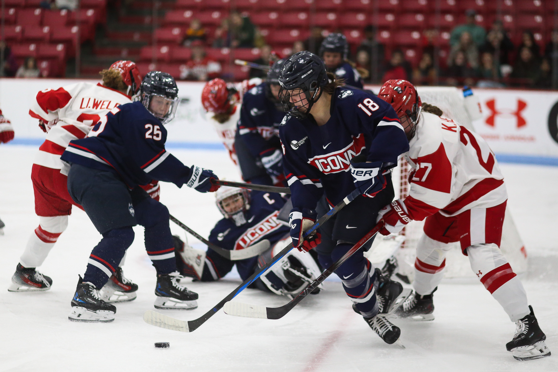Keila Healey (#27; BU) attempts to reach under Maya Serdachny (#18; UConn) to retrieve the puck back. Meanwhile, Jada Habisch (#25, UConn) looks to her right to see Serdachny is planning to pass the puck to her; February 22nd, 2025.