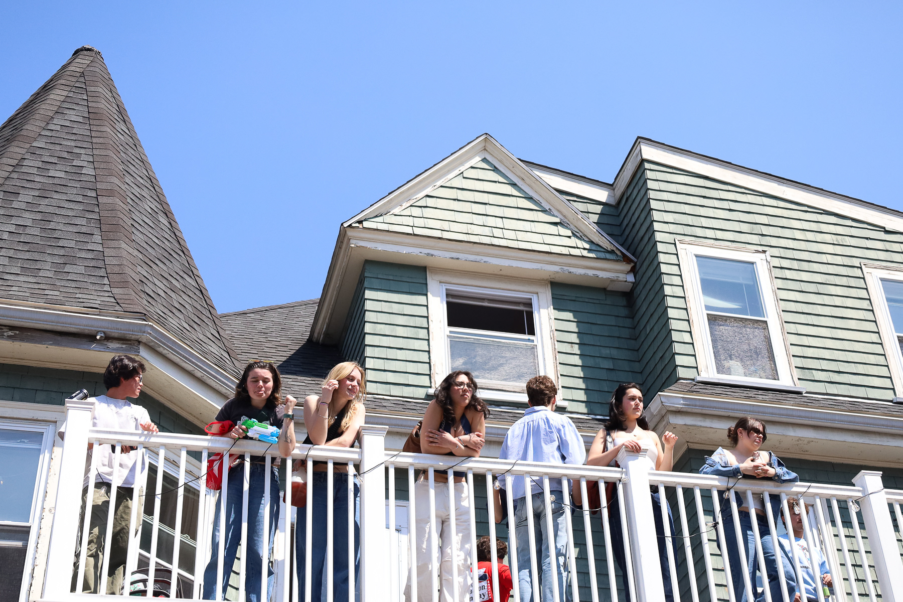 College students hang out on top of a balcony in an Allston frat house, spectating the large crowd on Marmon; April 21st, 2025.
