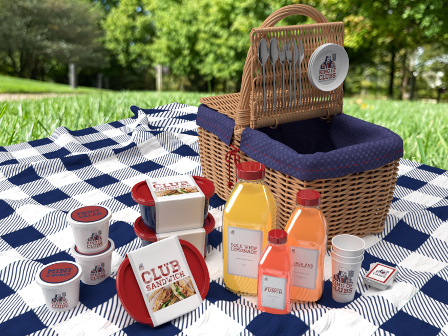 A picnic set, with different food items in their packaging and a basket, on a blanket with a park in the background.
