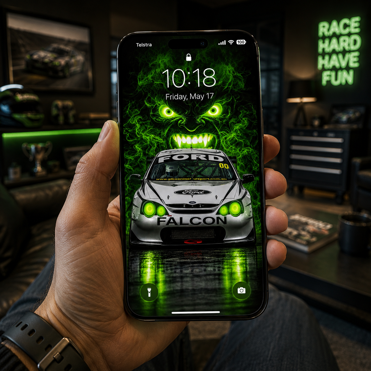 Phone-screen mock-up showing a man holding a smartphone displaying the Green Eyed Monster Ford Falcon automotive poster, featuring glowing green eyes and an illuminated race car.