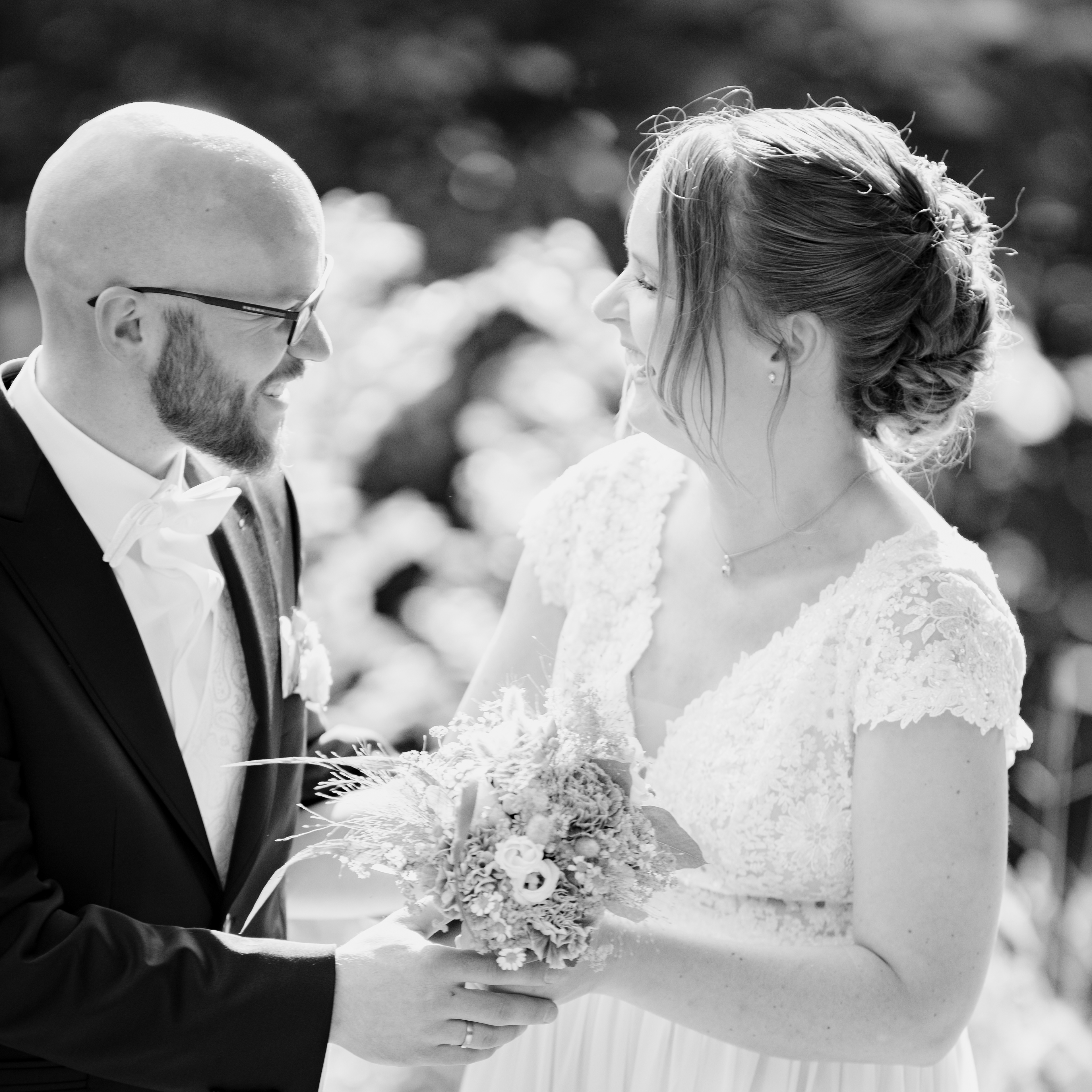 Brautpaar während des Brautpaarshootings. Die Sonne scheint von hinten und hüllt das Paar in einen Glanz. Beide halten den Brautstrauß gemeinsam fest und lachen sich herzlich an. das Bild ist in Schwarz Weiß 