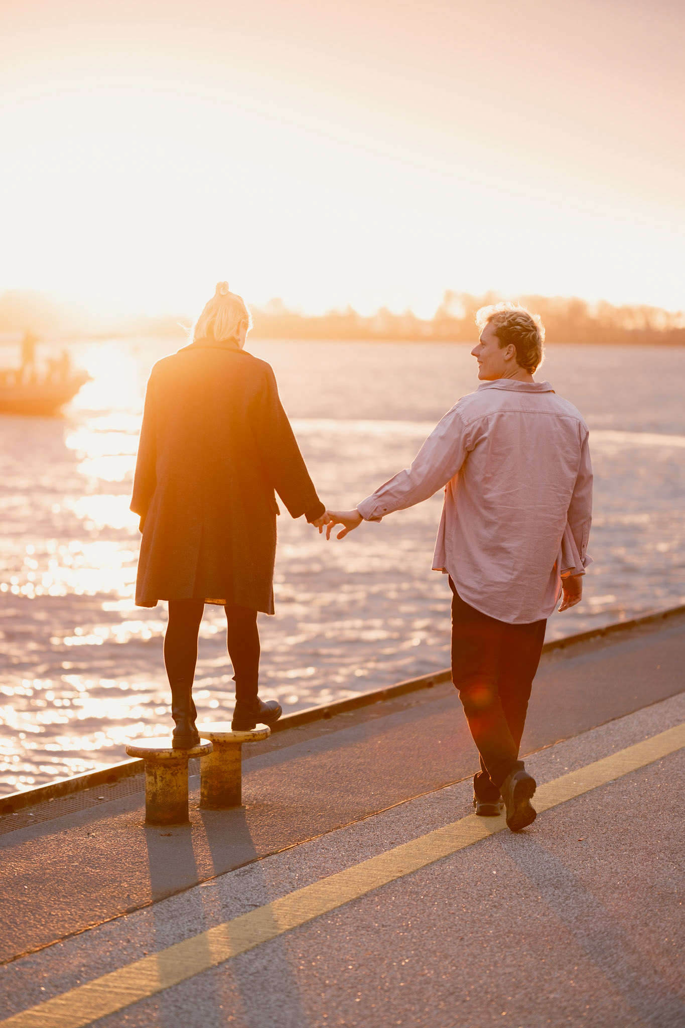 Verlobungsfotoshooting eines verlobten Paares am Elbstrand. Sie halten weich Händchen und spazieren am Fähranleger Övelgönne entlang. Die Sonne geht unter und er schaut sie verliebt an.
