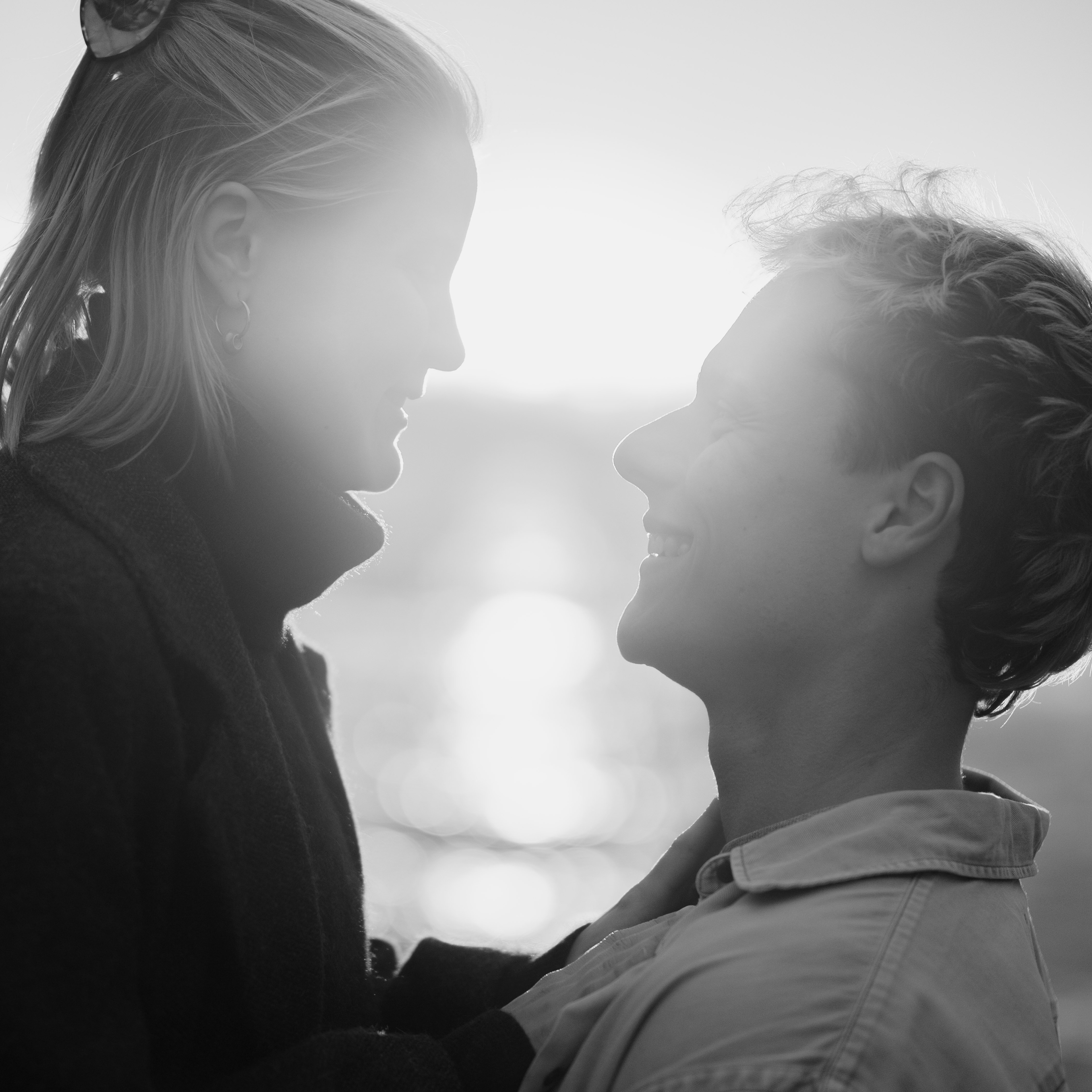 Verliebtes Paar steht vor dem Sonnenuntergang und lächelt sich an. Sie sind verlobt. Das Bild ist bei einem Verlobungsshooting entstanden und führt beim Klick zu der Experience, die von der Hochzeitsfotografie bei mir erwartet werden kann. Das Bild ist in Schwarz Weiß.