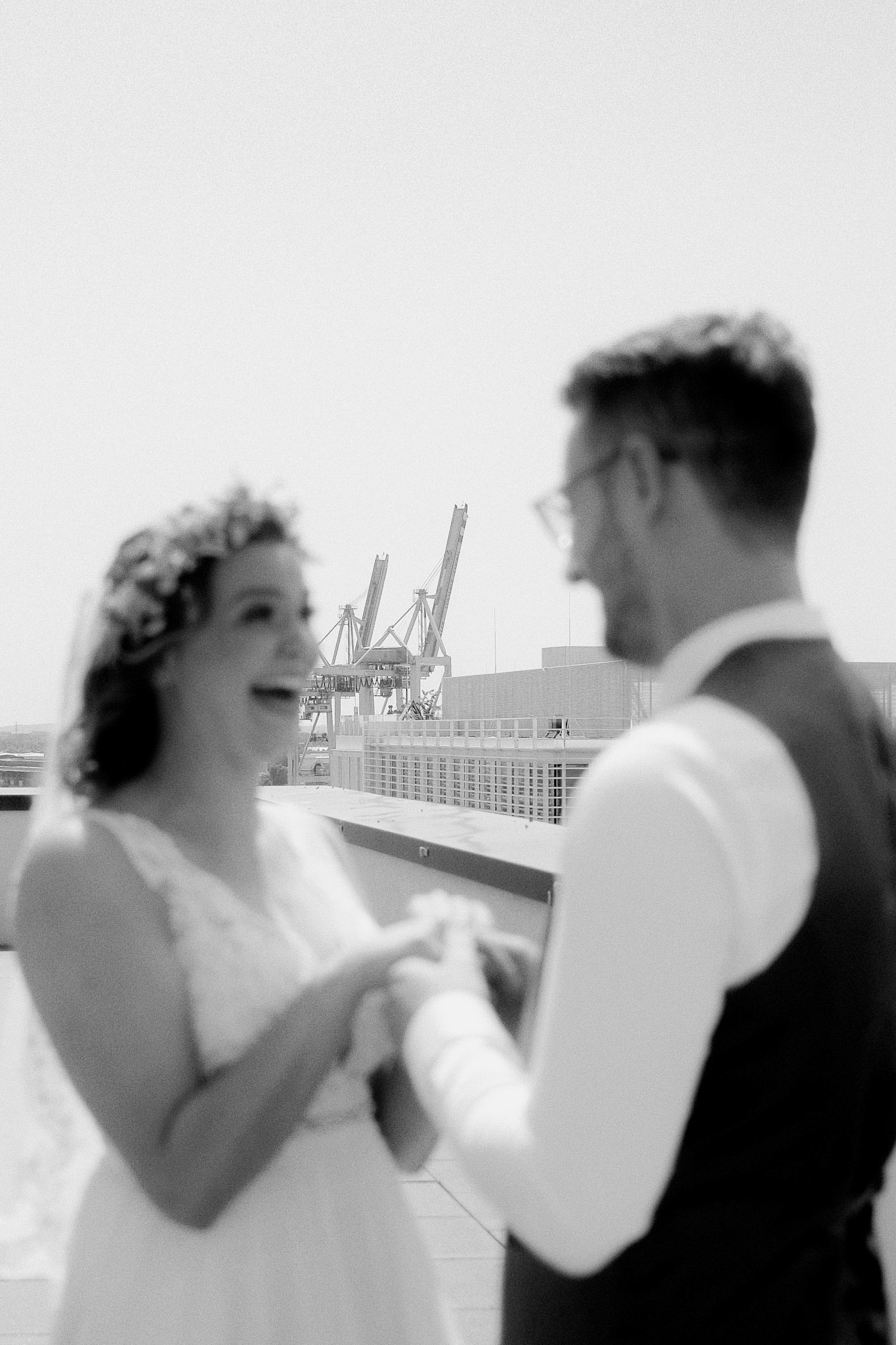 Brautpaar sieht sich zum ersten mal und lacht sich an. Im Hintergrund ist der Hamburger Hafen zu sehen. Das Bild entstand beim First Look auf einer Dachterrasse. Das Brautpaar ist in künstlerischer Unschärfe, man erkennt aber wie sehr sie sich freuen. Sie betrachten sich.