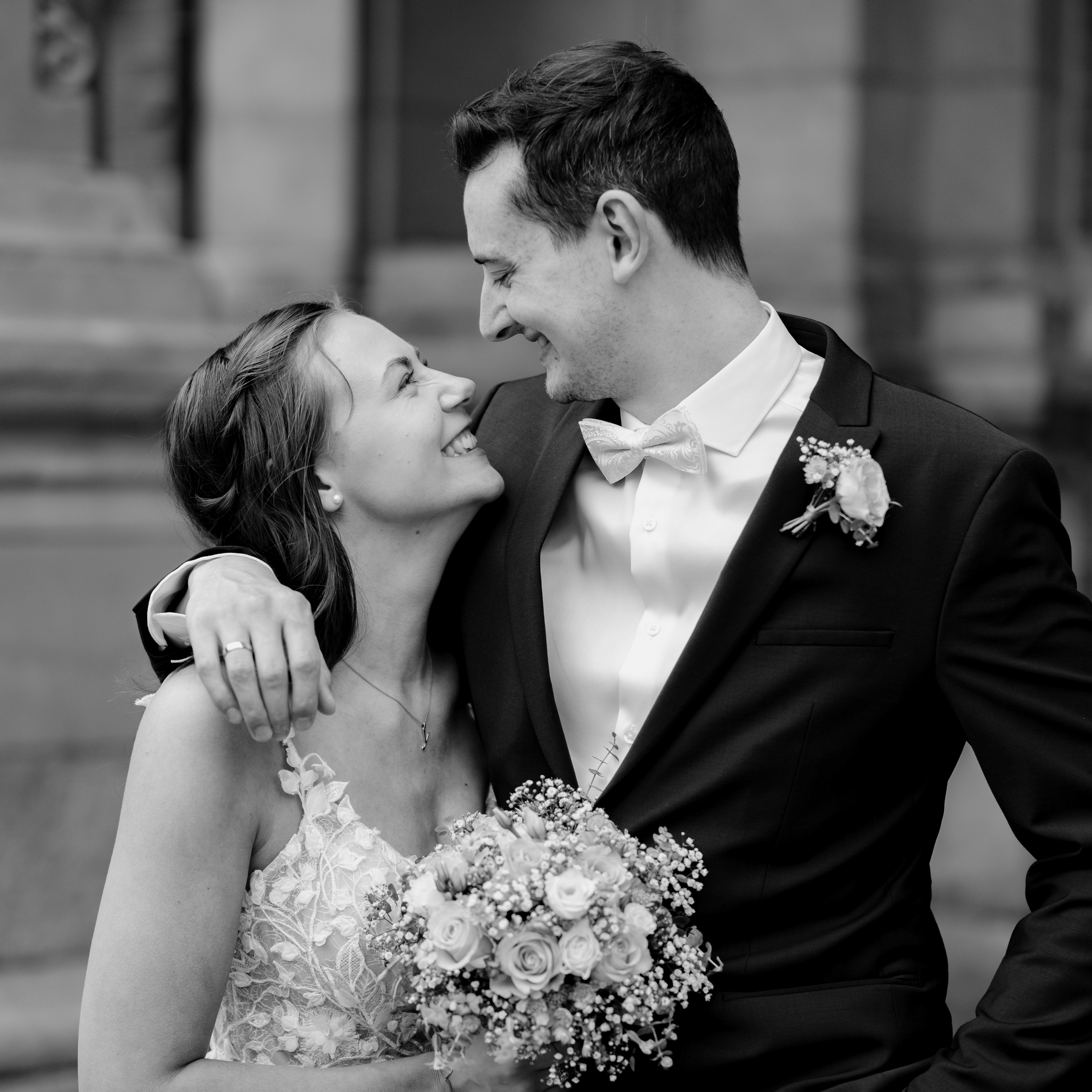 ein Frisch vermähltes Paar steht vor dem Harburger Rathaus und lächelt sich strahlend an. Er hat seinen Arm um sie gelegt. Sie hält den Brautstrauß fest. Man sieht seinen Ehering. Beide sehen super glücklich aus. Das Bild ist in Schwarz Weiß.