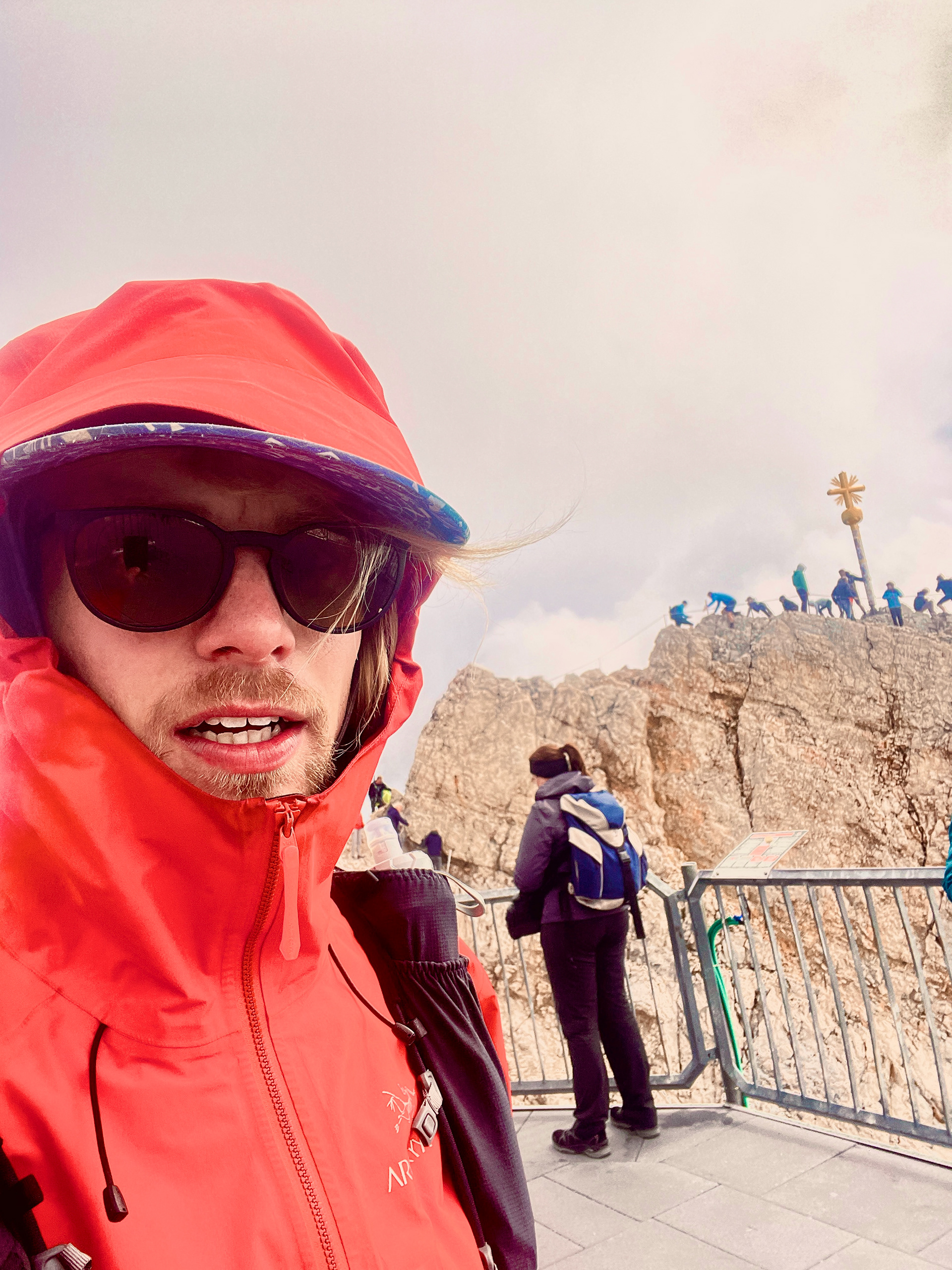 Silas steht an der Spitze der Zugspitze nachdem er auf den Berg gelaufen ist. Er trägt eine Sonnenbrille und eine Hardshell Jacke. Das Wetter scheint nicht sehr gemütlich zu sein. Er muss sich beeilen wieder vom Berg runter zu kommen.