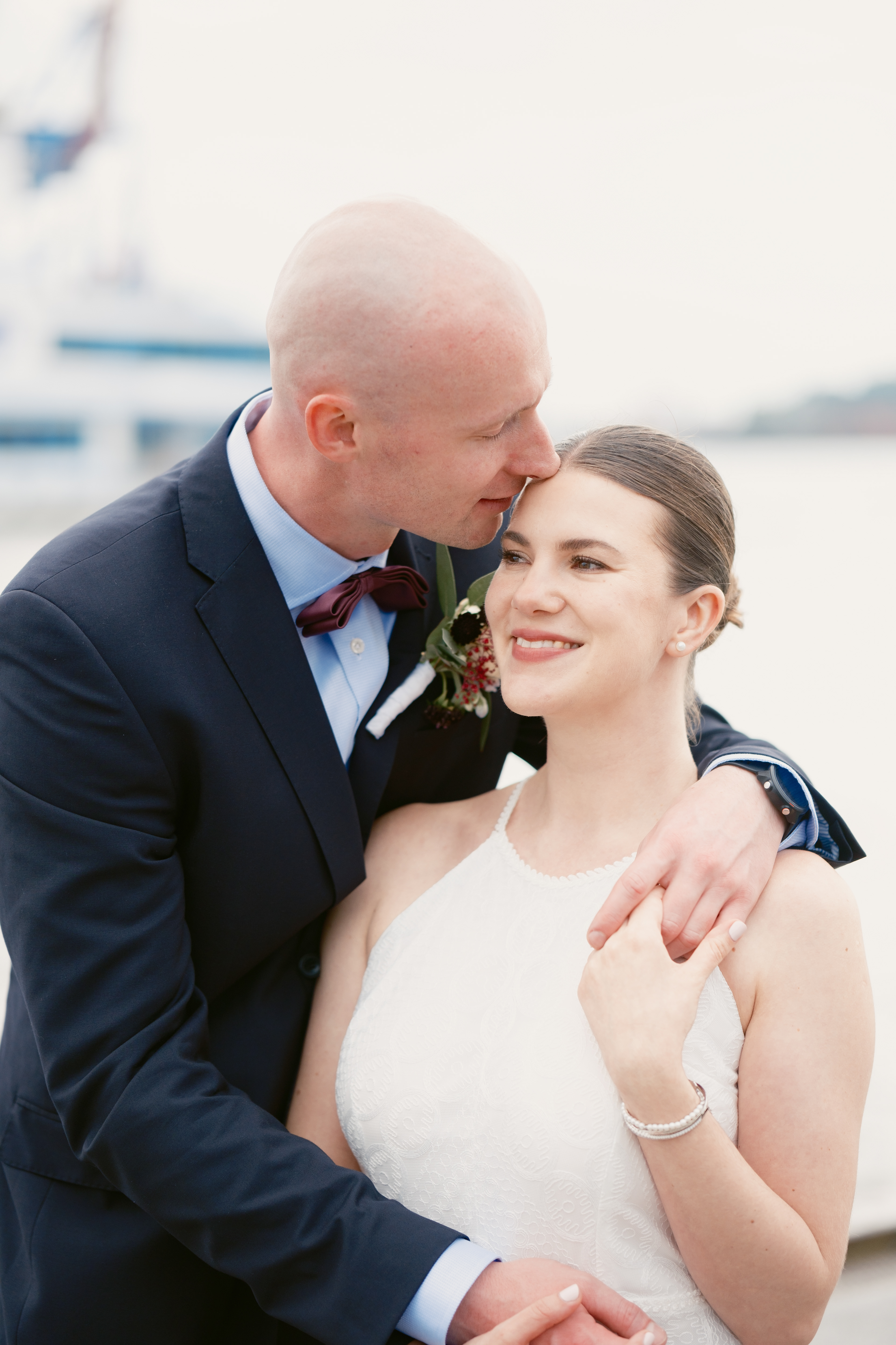 Brautpaar steht am Elbstrand in inniger Umarmung und er küsst sie sanft auf die Stirn. Beide lächeln vor Freude. Das Licht ist sanft und weich. 