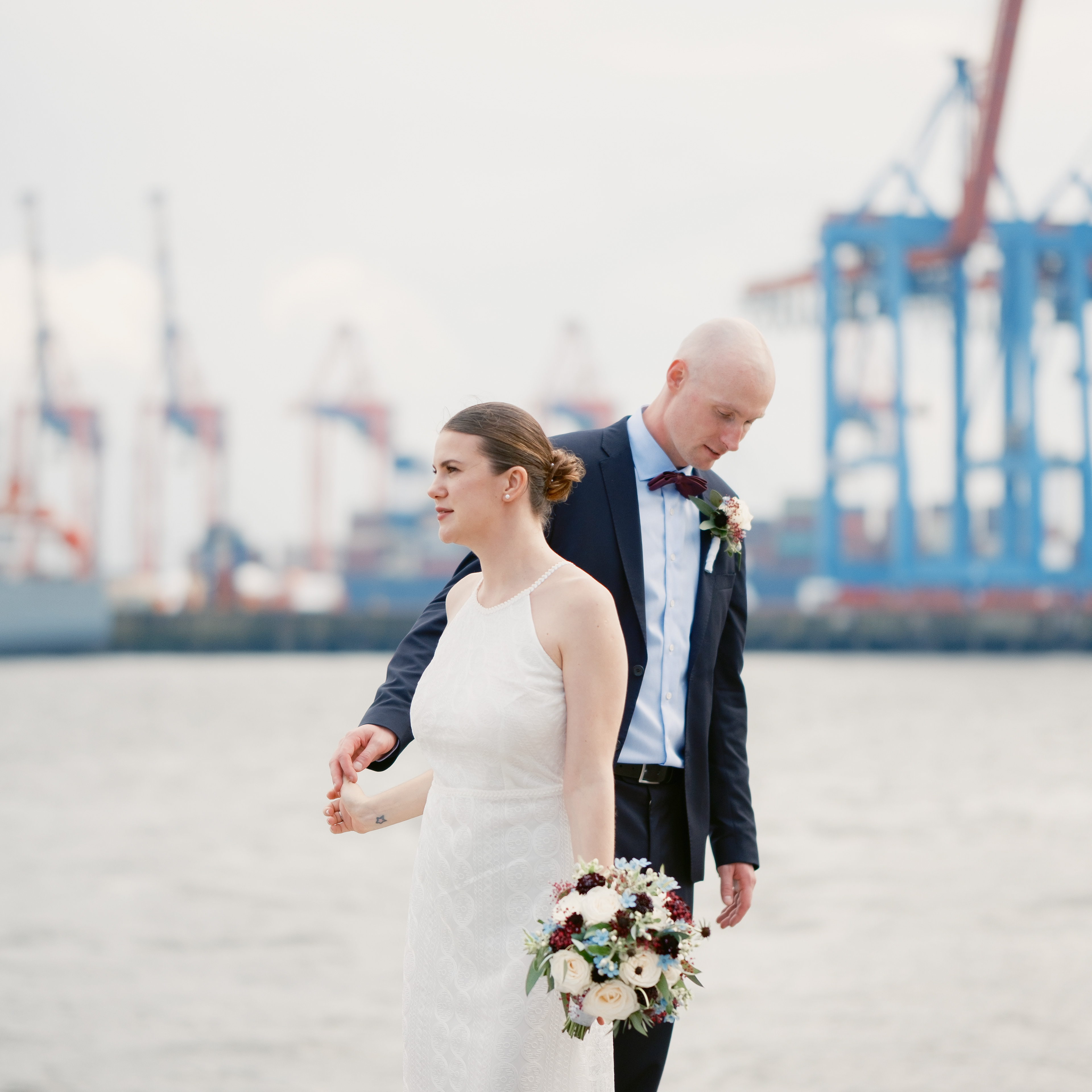 Frisch vermähltes Brautpaar steht am Elbstrand und hält sanft Händchen. Sie geht an ihm vorbei. er schaut weich auf den Boden. Beide sehen sehr edel aus. Im Hintergrund sieht man die Kräne des Hamburger Hafens. Das Licht ist weich, warm und angenehm.