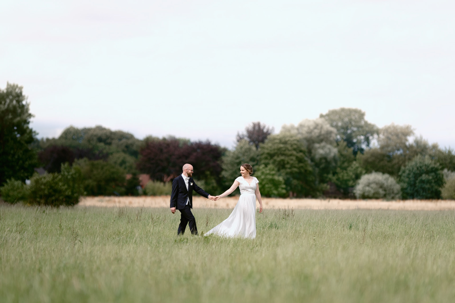 Hochzeitspaar geht über ein weites Feld. Sie halten Händchen und beschreiten beide gemeinsam den Weg der Liebe. Sie schaut über die Schulter zu ihm zurück.
