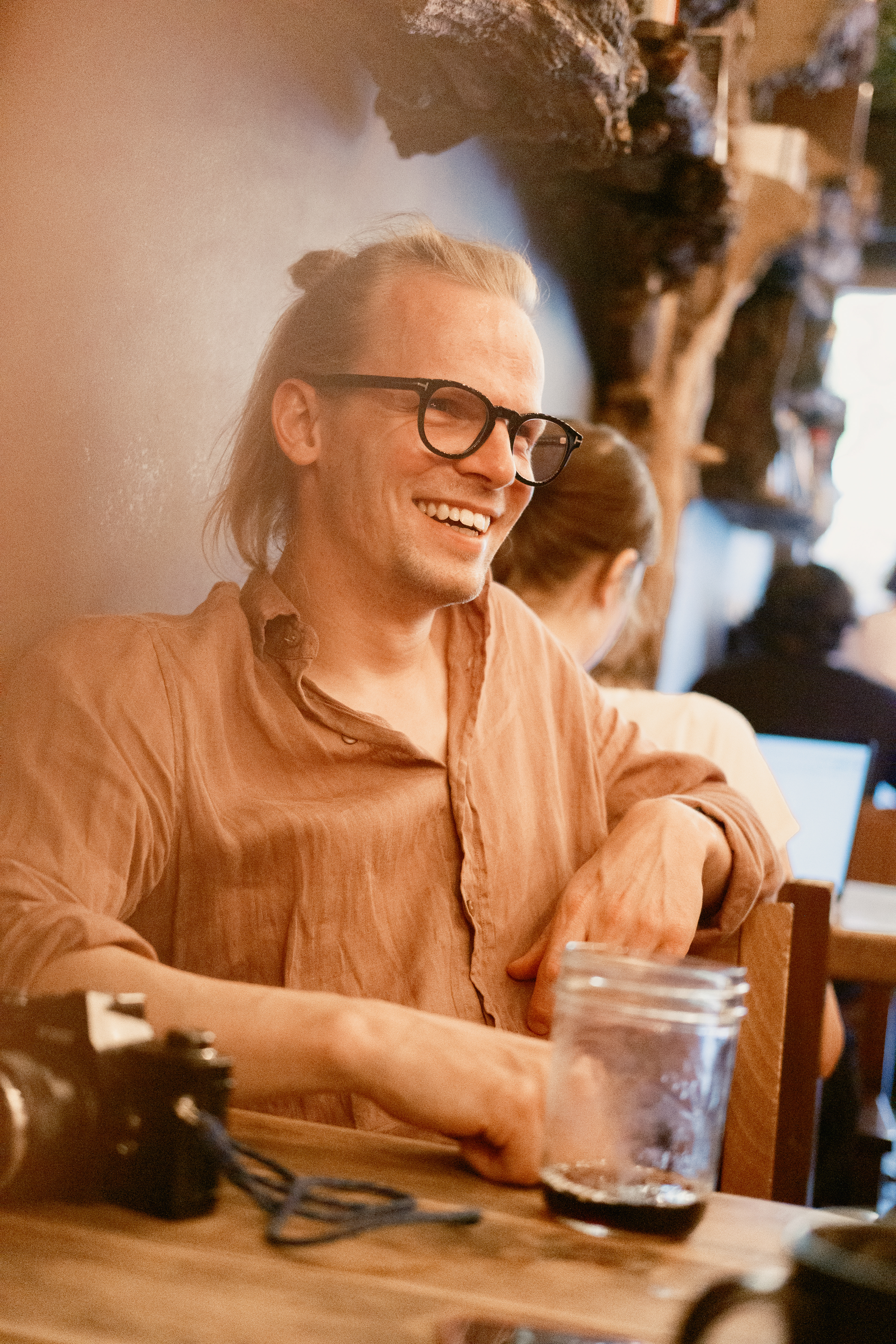 Silas Wagner. Fotograf aus deiner Nähe. Er sitzt in einem Kaffee mit seiner alten analogen Kamera vor sich auf dem Tisch. Er lächelt sympathisch und trägt ein lockeres Leinenhemd. 
