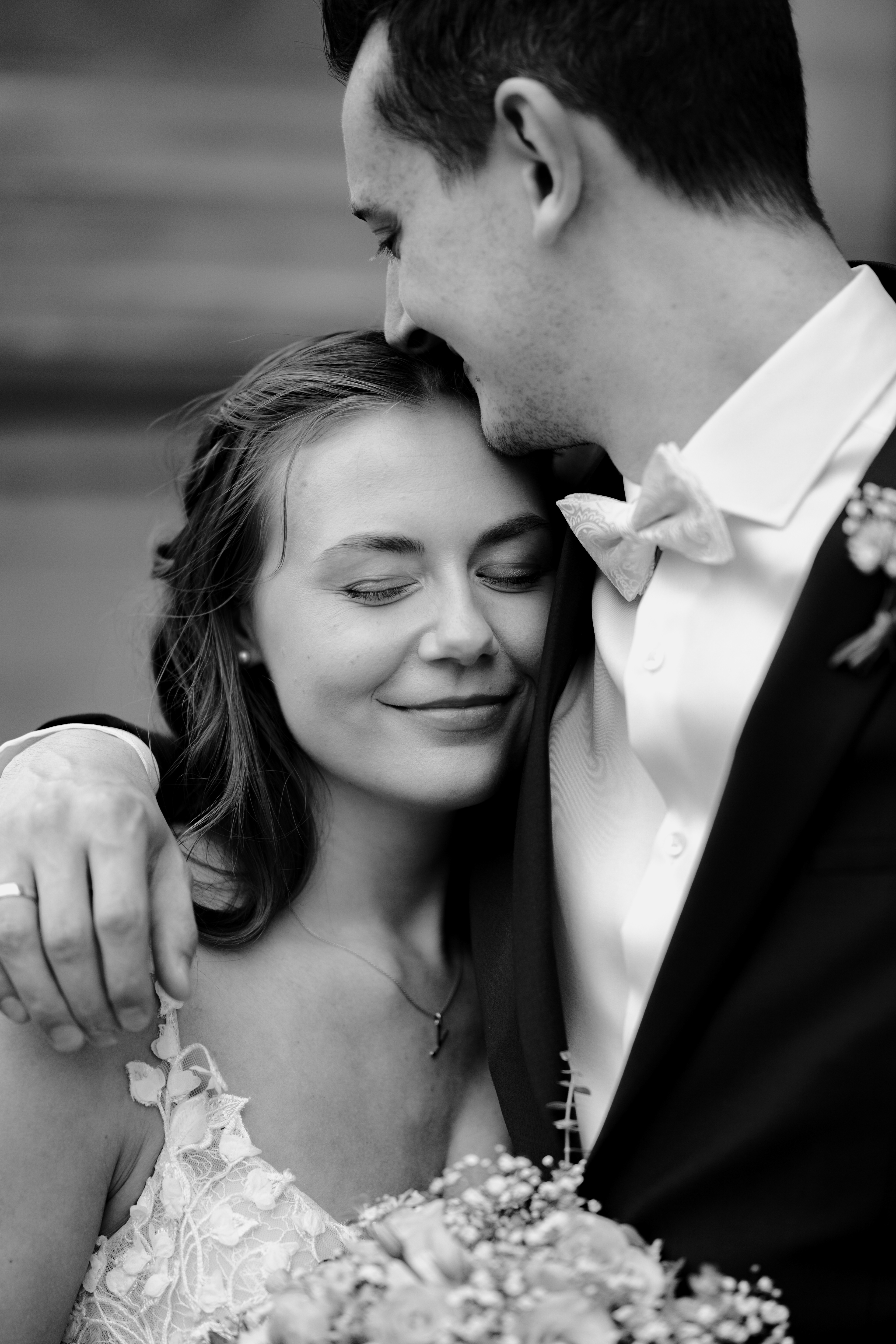 Brautpaar steht vor dem Standesamt. Sie ist an seine Schulter gekuschelt und er küsst sie auf die Stirn. Man sieht seinen Ehering. Sie lächelt mit geschlossenen Augen in sich rein. Das Bild in in Schwarz weiß.