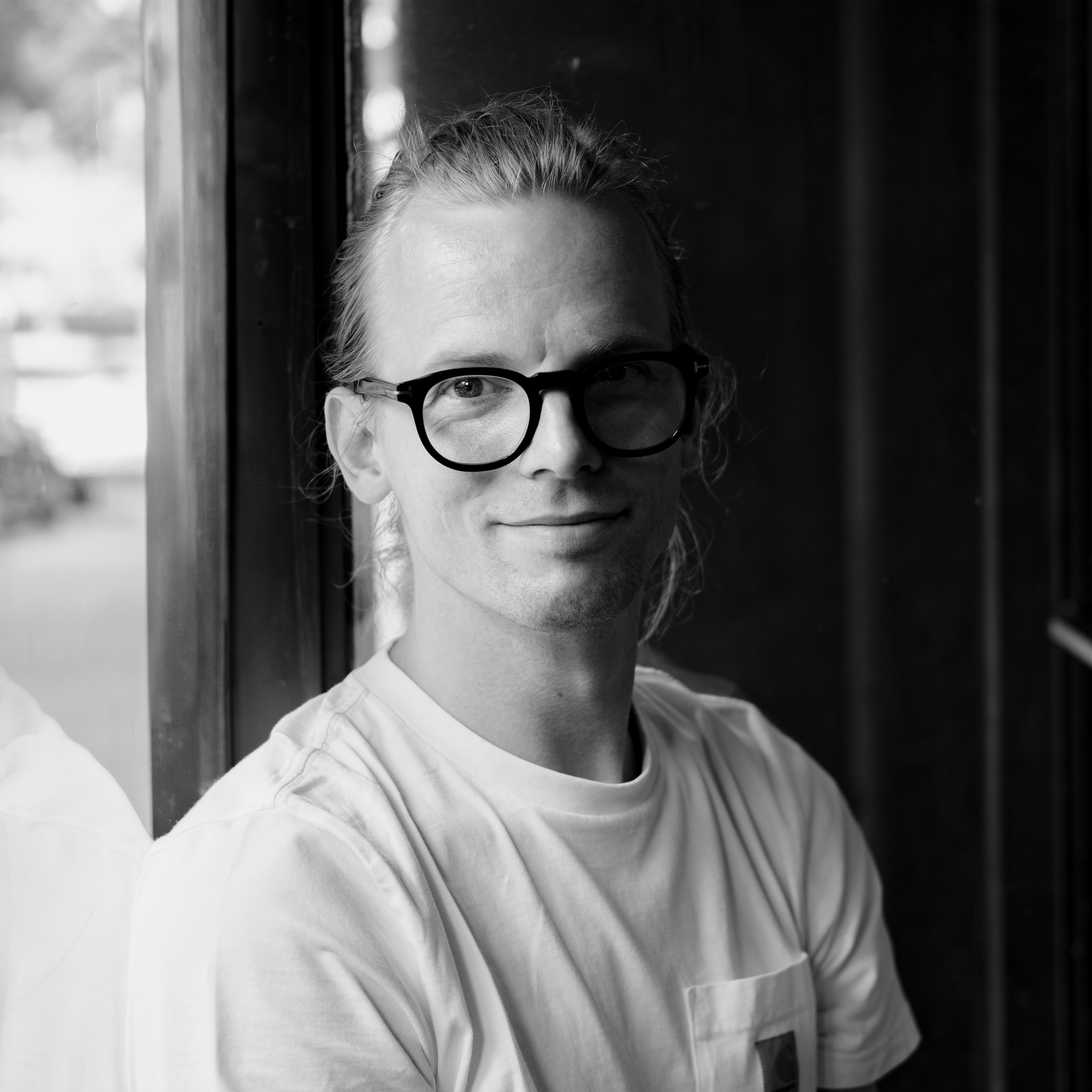 Silas Wagner, dein Fotograf aus deiner Nähe. Er sitzt an einem Fenster und lächelt weich direkt in die Kamera. Er trägt eine große schwarze Brille und hat zurückgebundene Haare. Das Bild ist in schwarz weiß.