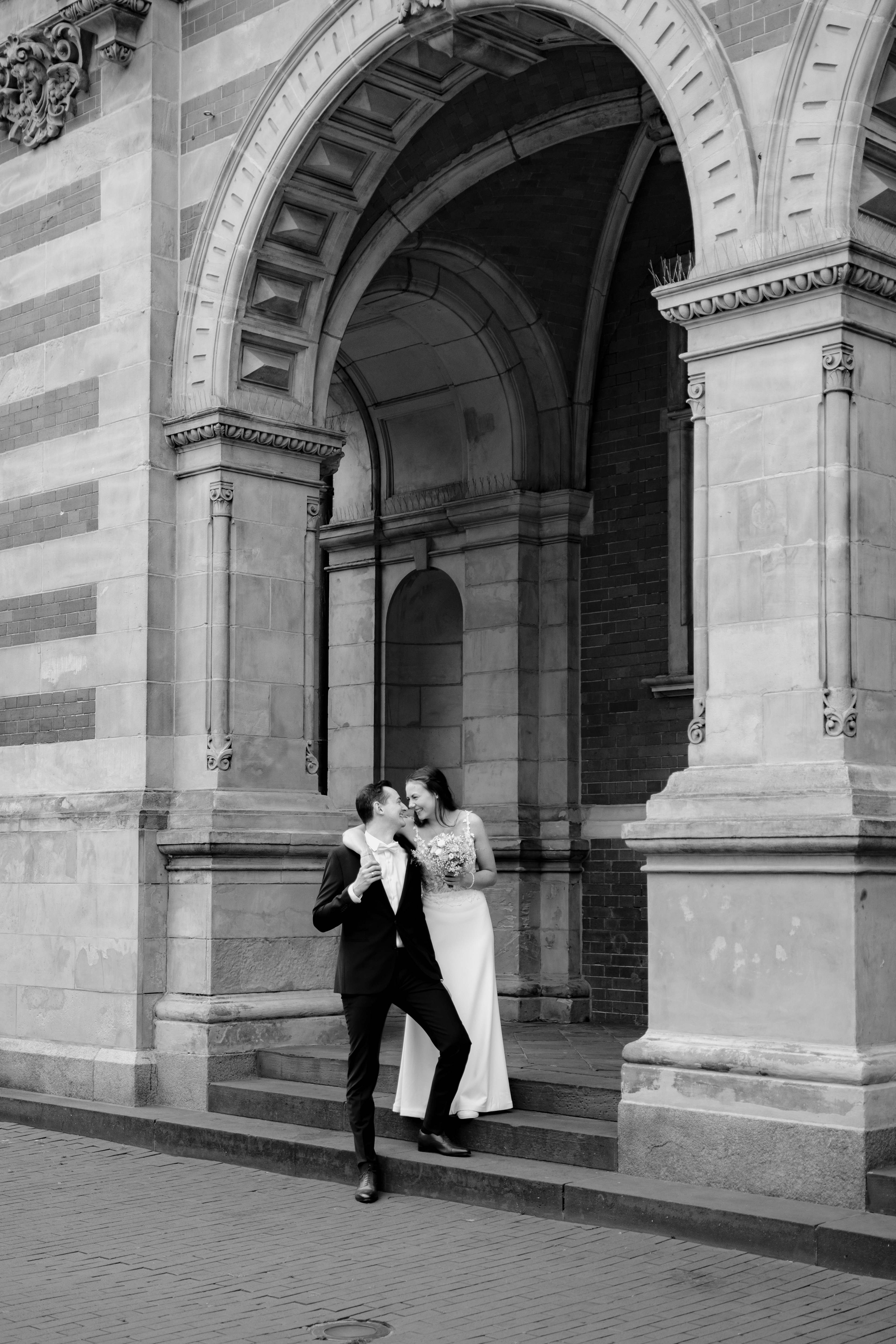 Frisch vermähltes Paar steht vor dem Harburger Rathaus auf der Treppe und schaut sich an. Das Bild ist in schwarz weiß. Es sieht monumental aus durch die hohen Säulen und das weiche Licht. Sie steht leicht über ihm und schlingt ihren Arm um ihn. 
