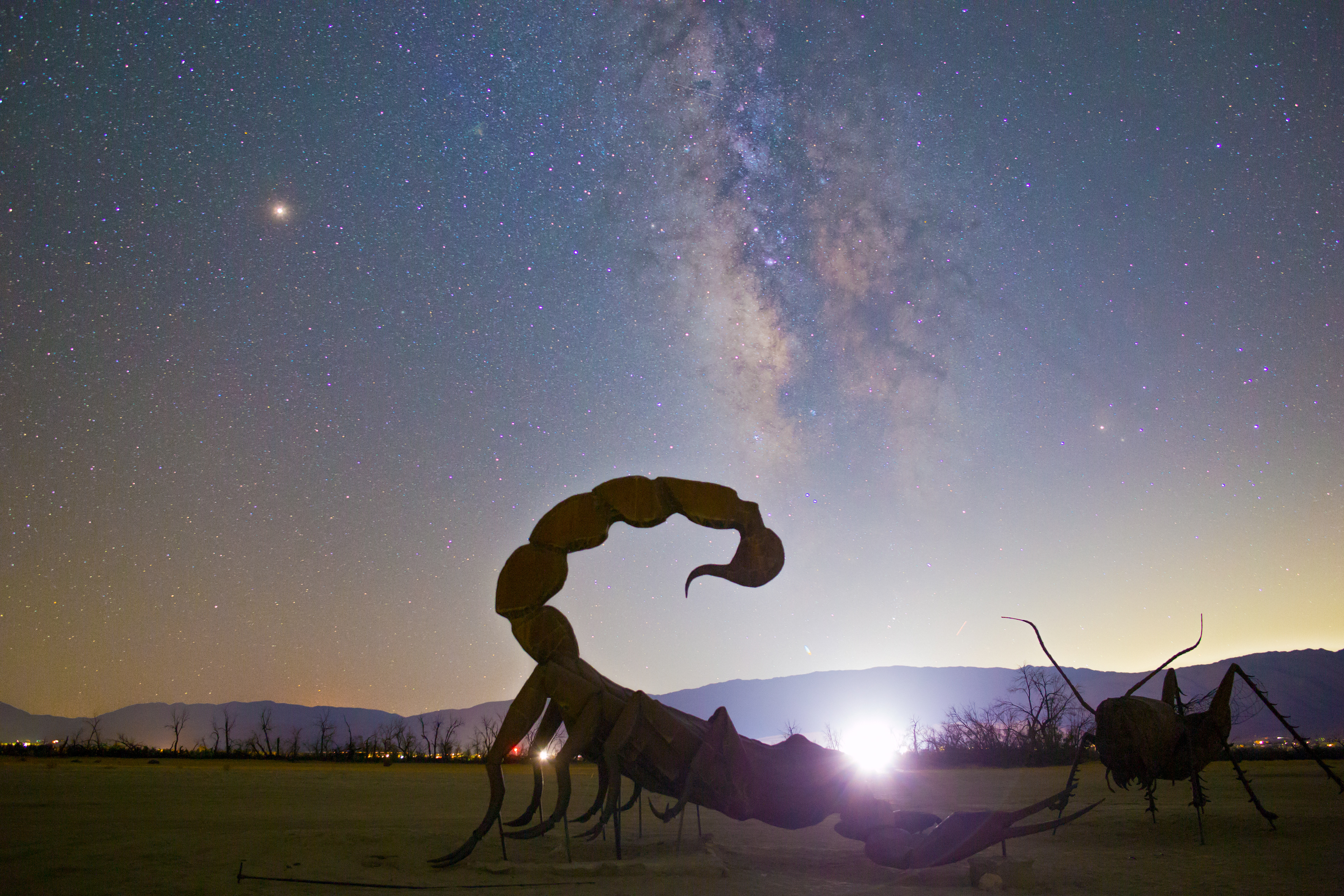A scorpion statue is lit up by Borrego Springs in front of Mars and the Milky Way.