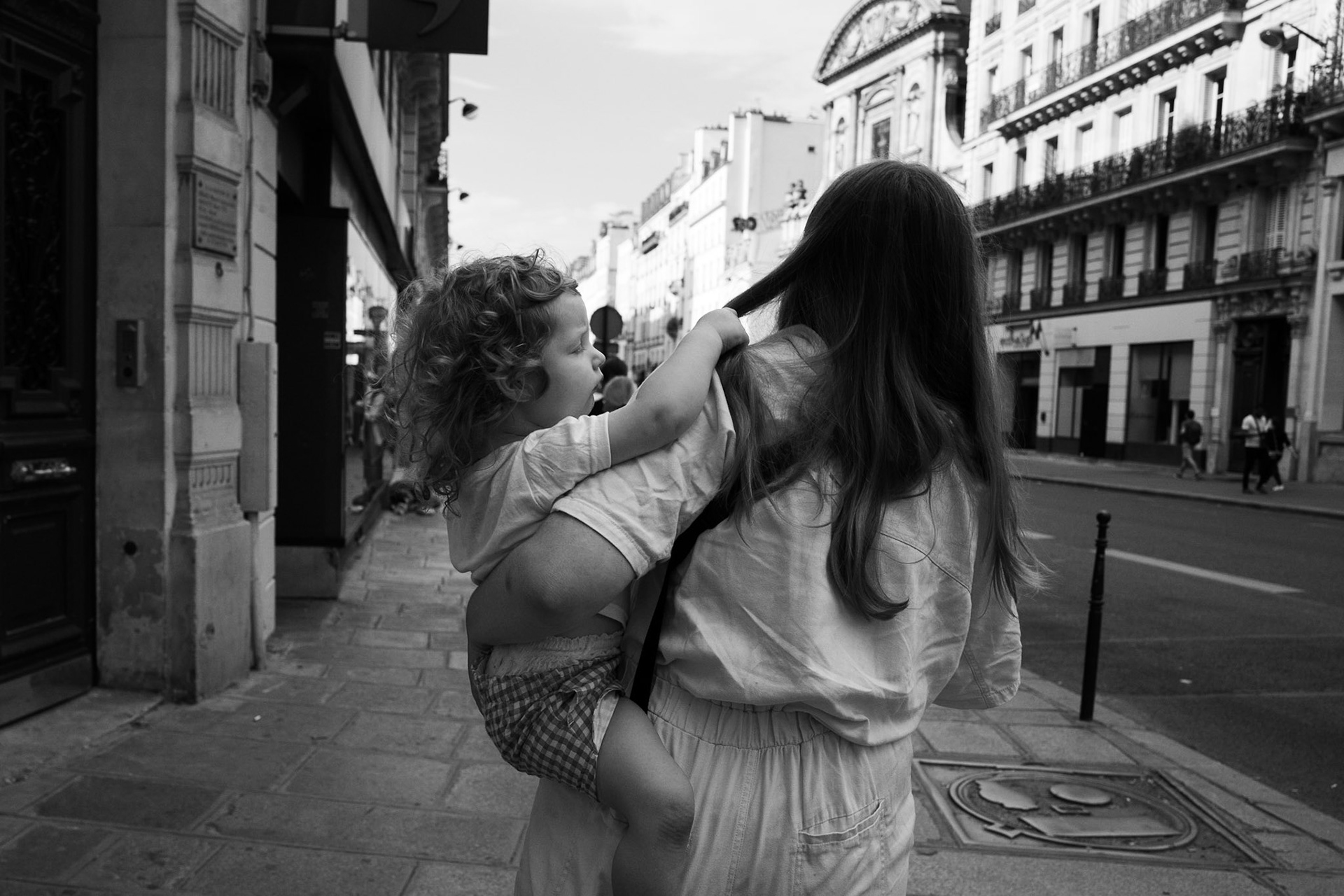 Une femme portant un enfant dans les bras marche sur un trottoir haussmannien. L’enfant s’accroche à une mèche de cheveux, regardant la ville avec curiosité.