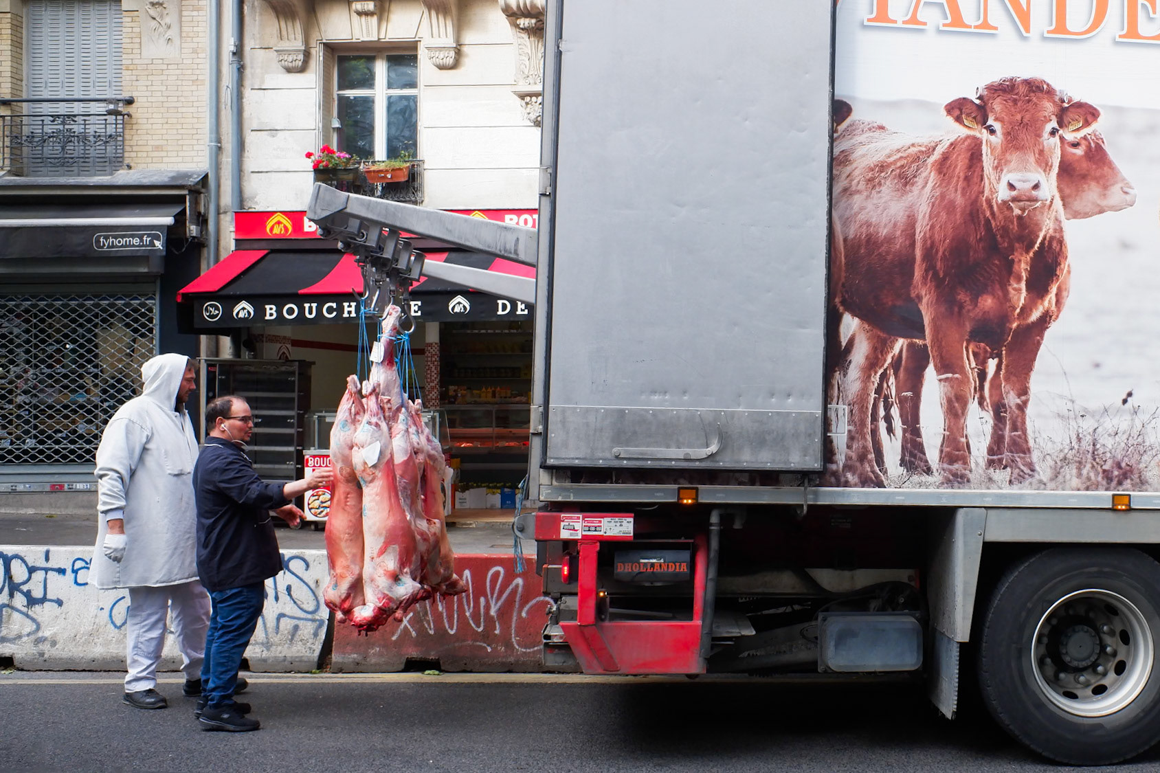 Déchargement de carcasses dans la rue devant une boucherie, avec un camion illustré d’un troupeau de vaches vivantes — un jeu visuel sur la chaîne alimentaire.
