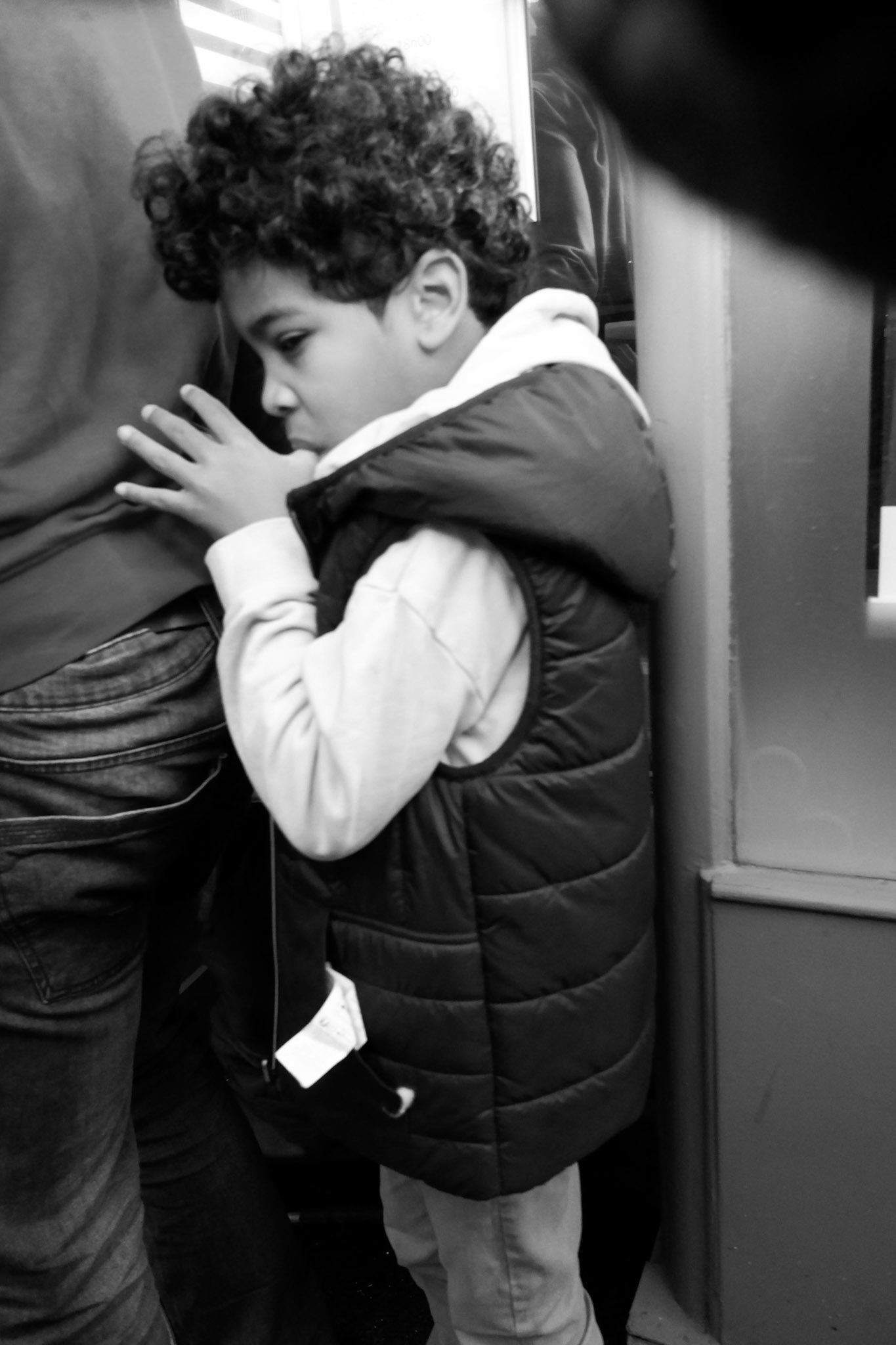 Un jeune garçon recroquevillé dans un coin du métro, le regard pensif, les doigts dans la bouche, blotti contre un adulte — scène intime et silencieuse dans la foule urbaine.