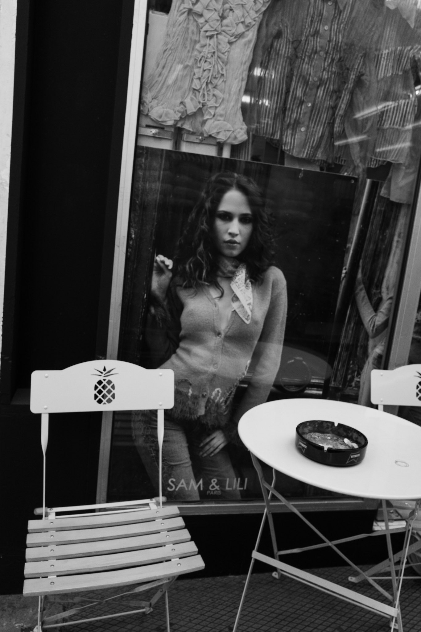 Affiche de mode en vitrine montrant une femme regardant fixement le photographe, partiellement cachée derrière un mobilier de terrasse avec cendrier.