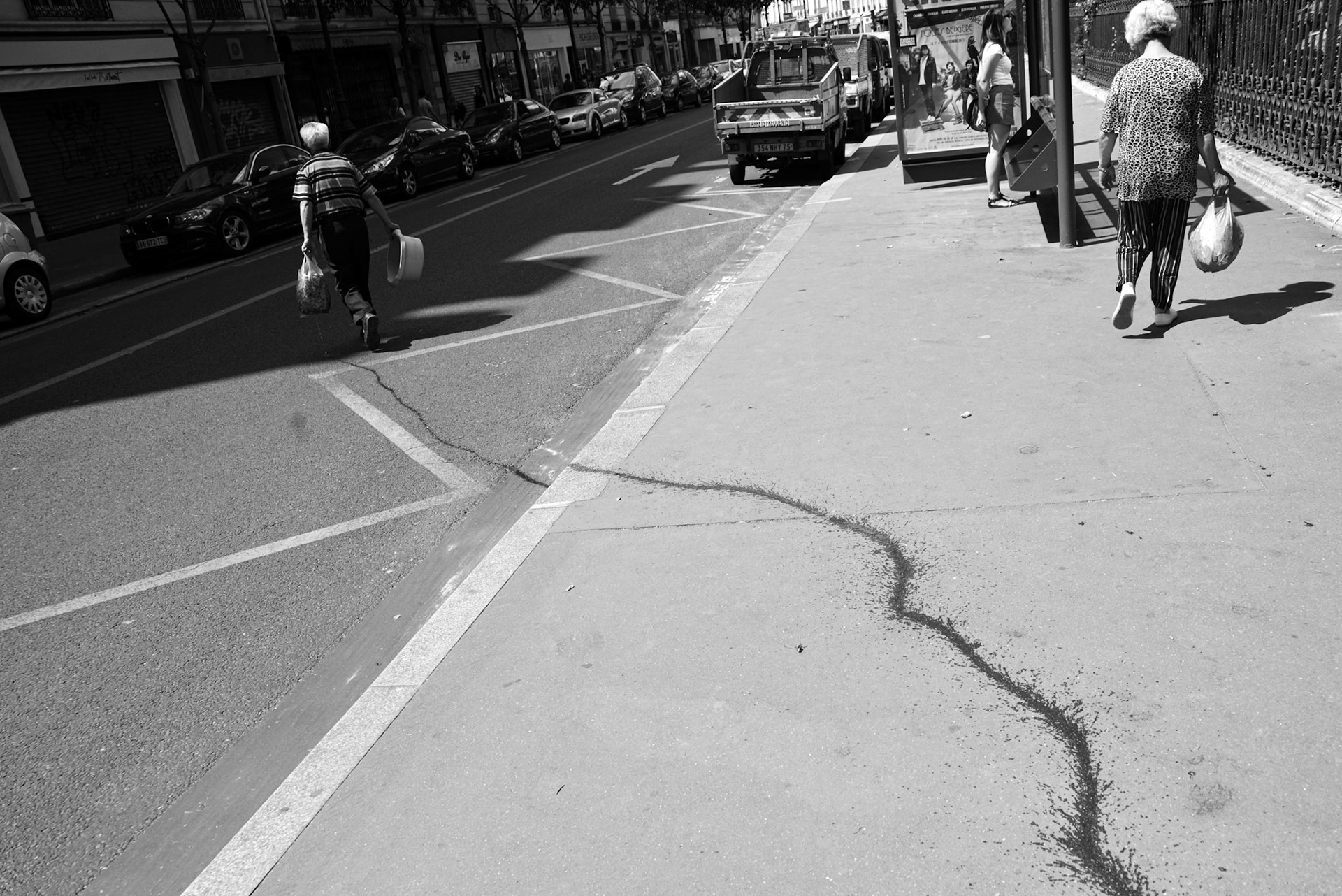 Rue ensoleillée avec un homme et un femmes marchant en sens opposé, séparées par une longue traînée noire serpentant sur le trottoir.